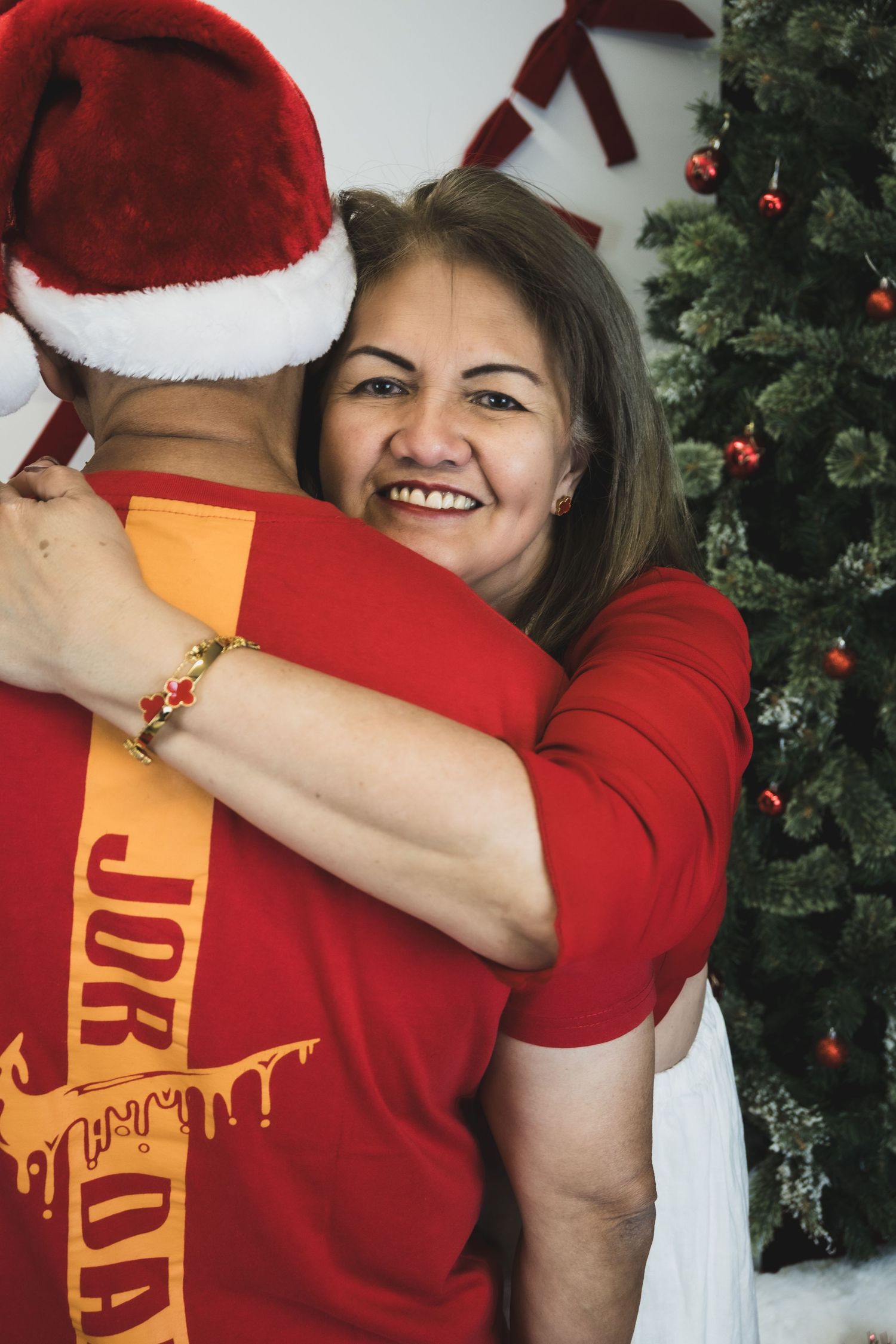 Woman in red shirt smiling while hugging person in Santa hat near decorated Christmas tree.