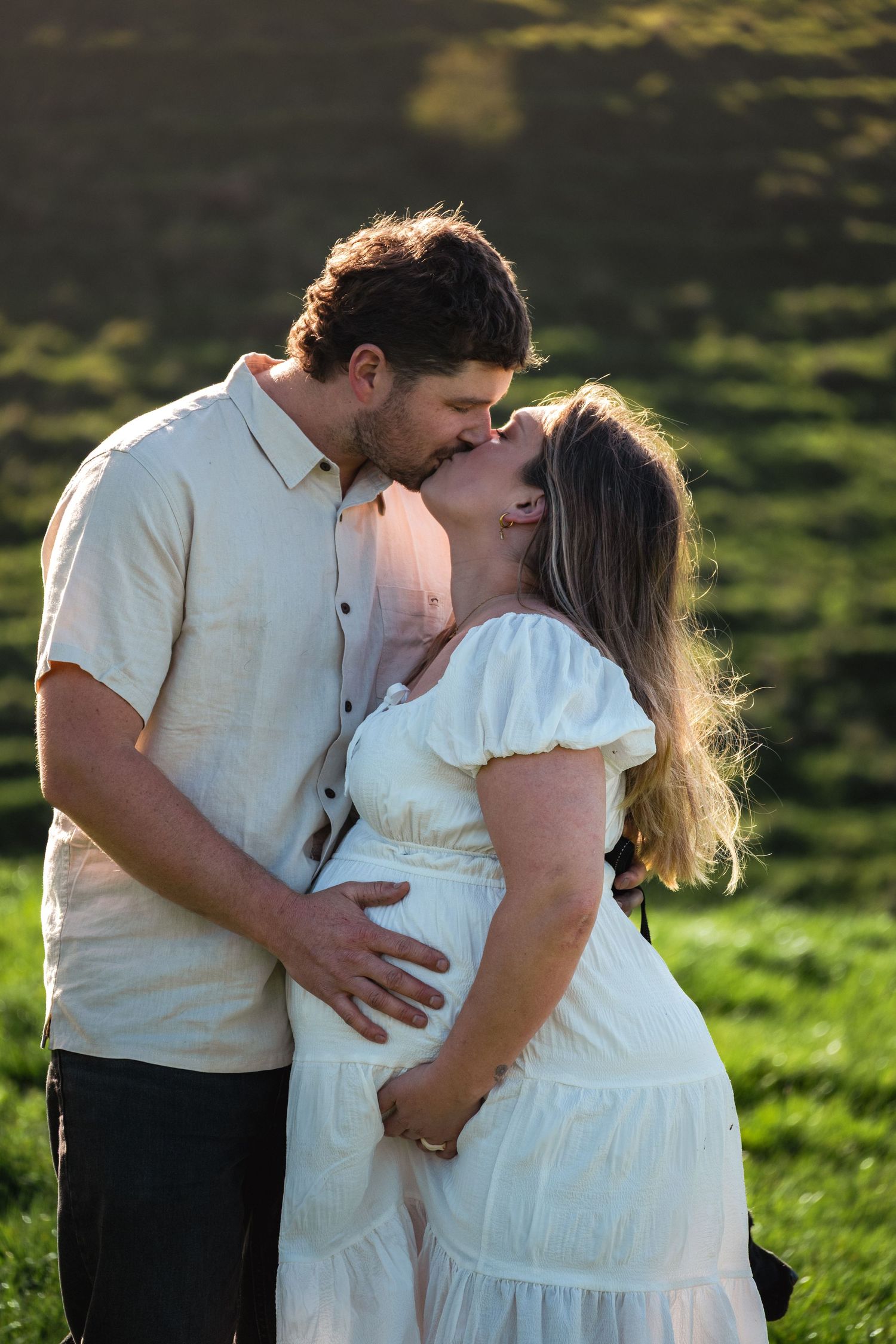 Pregnant couple sharing a tender kiss outdoors, woman in white dress cradling baby bump, man in beige shirt, green hills background.