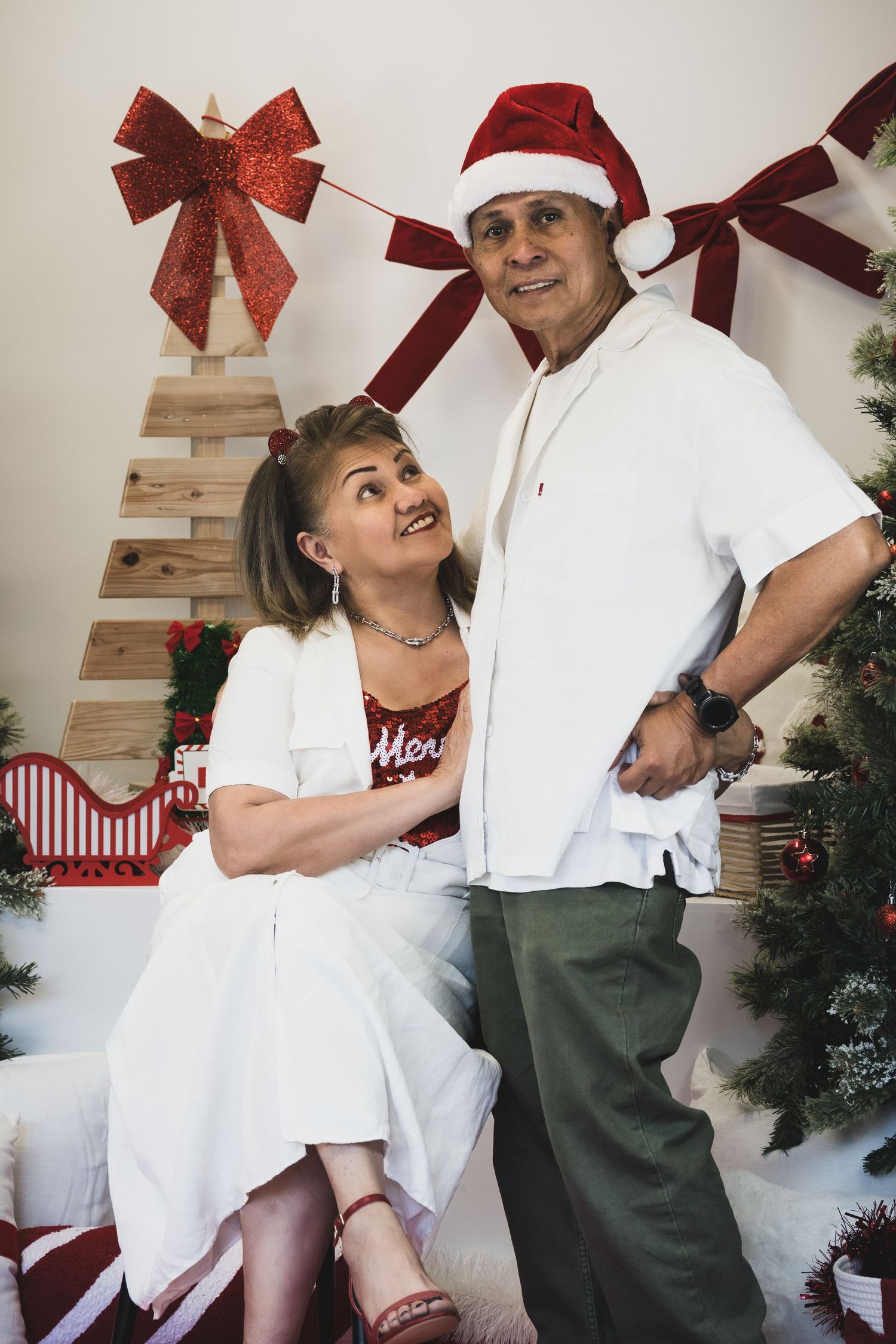 Elderly couple posing together in Christmas setting with Santa hat, decorated tree, and festive red bow backdrop.