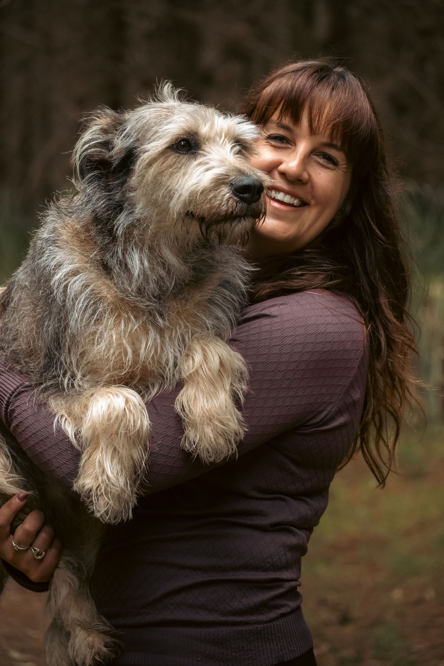 Smiling woman with bangs holding a large fluffy gray terrier mix dog outdoors in a wooded setting.