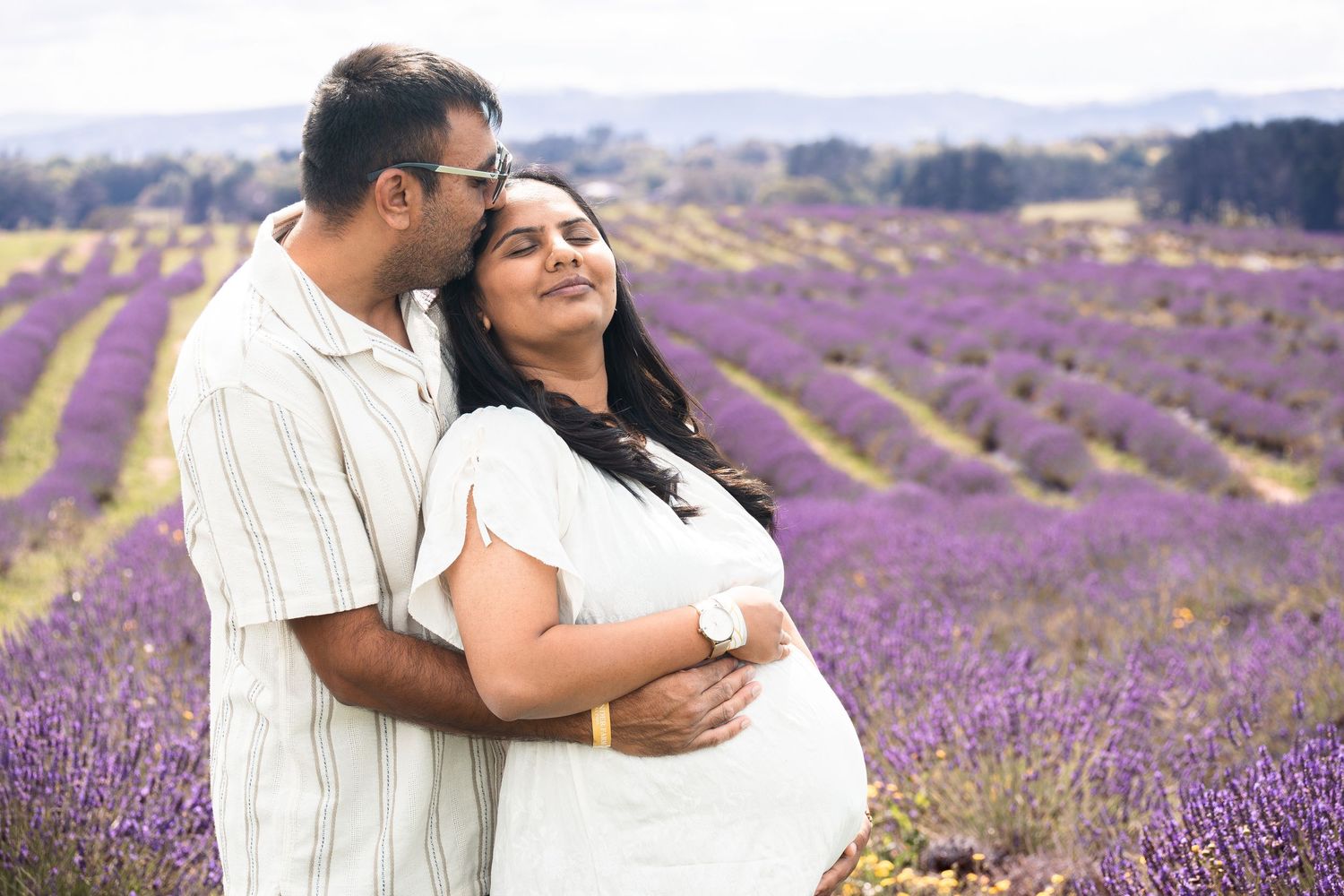 Pregnant couple embracing in a blooming lavender field, woman in white dress, man in striped shirt, outdoors.