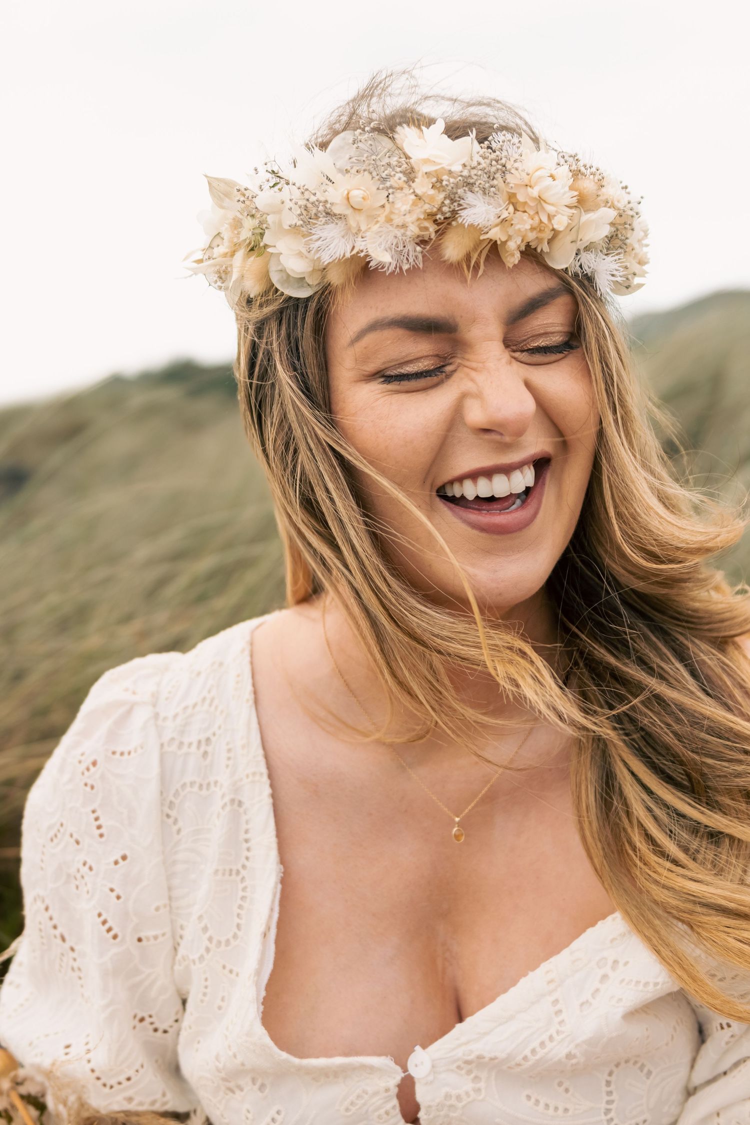 Laughing woman wearing a boho floral crown and white dress outdoors with green hills in background.