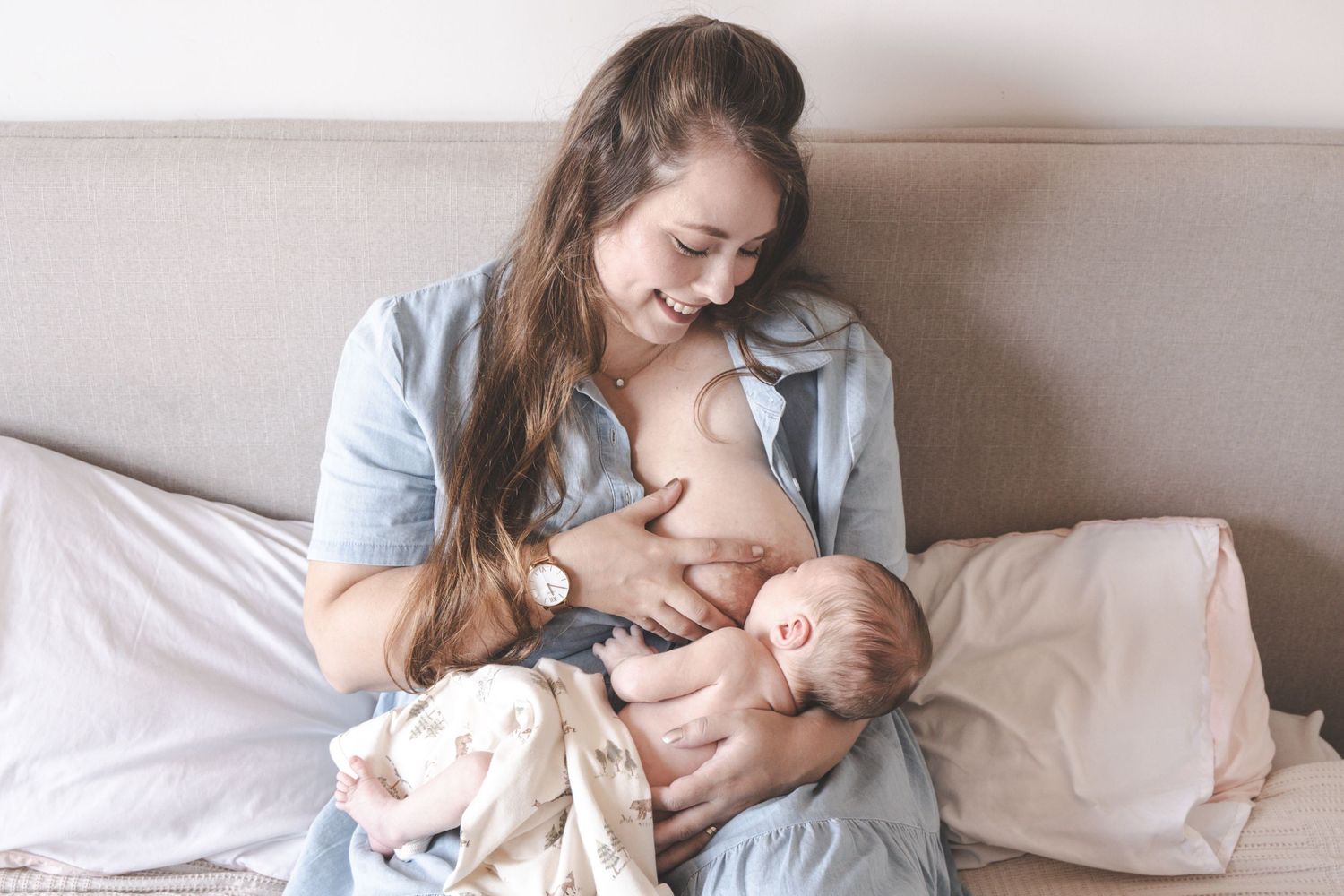 Young mother breastfeeding newborn baby while sitting on bed, smiling warmly in soft natural light.