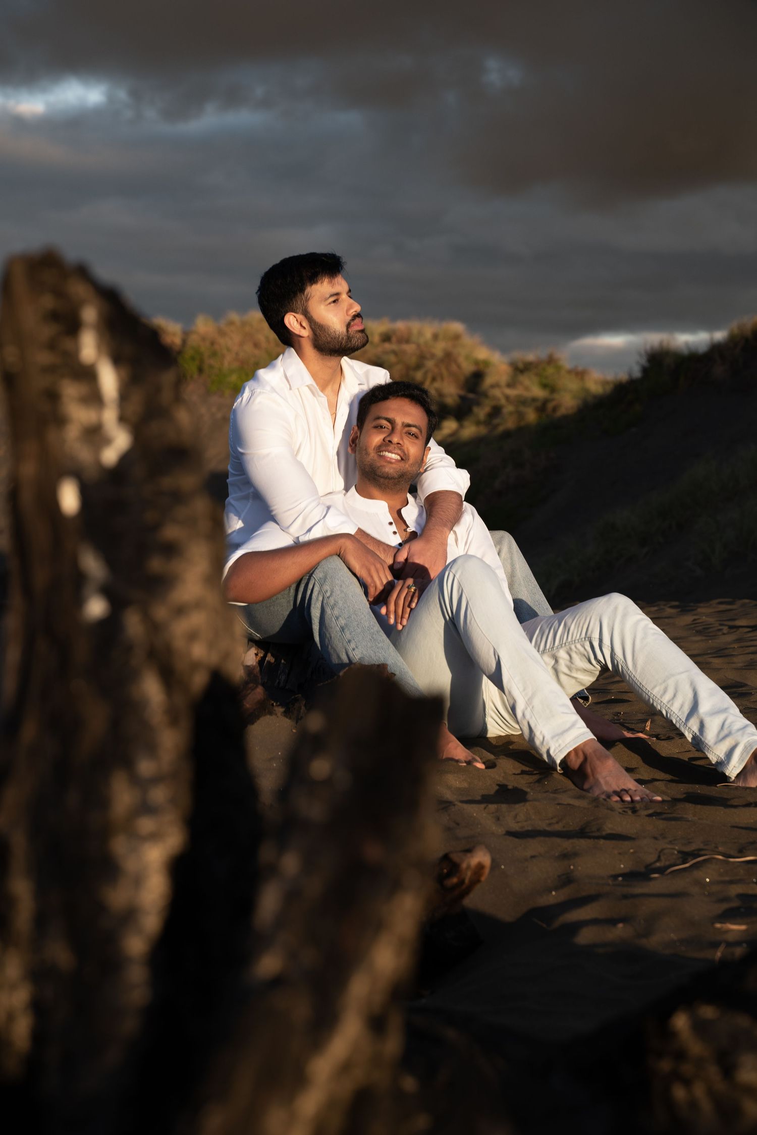 Two men in white shirts sit together on a sandy beach at sunset, sharing a tender embrace under dramatic cloudy skies.