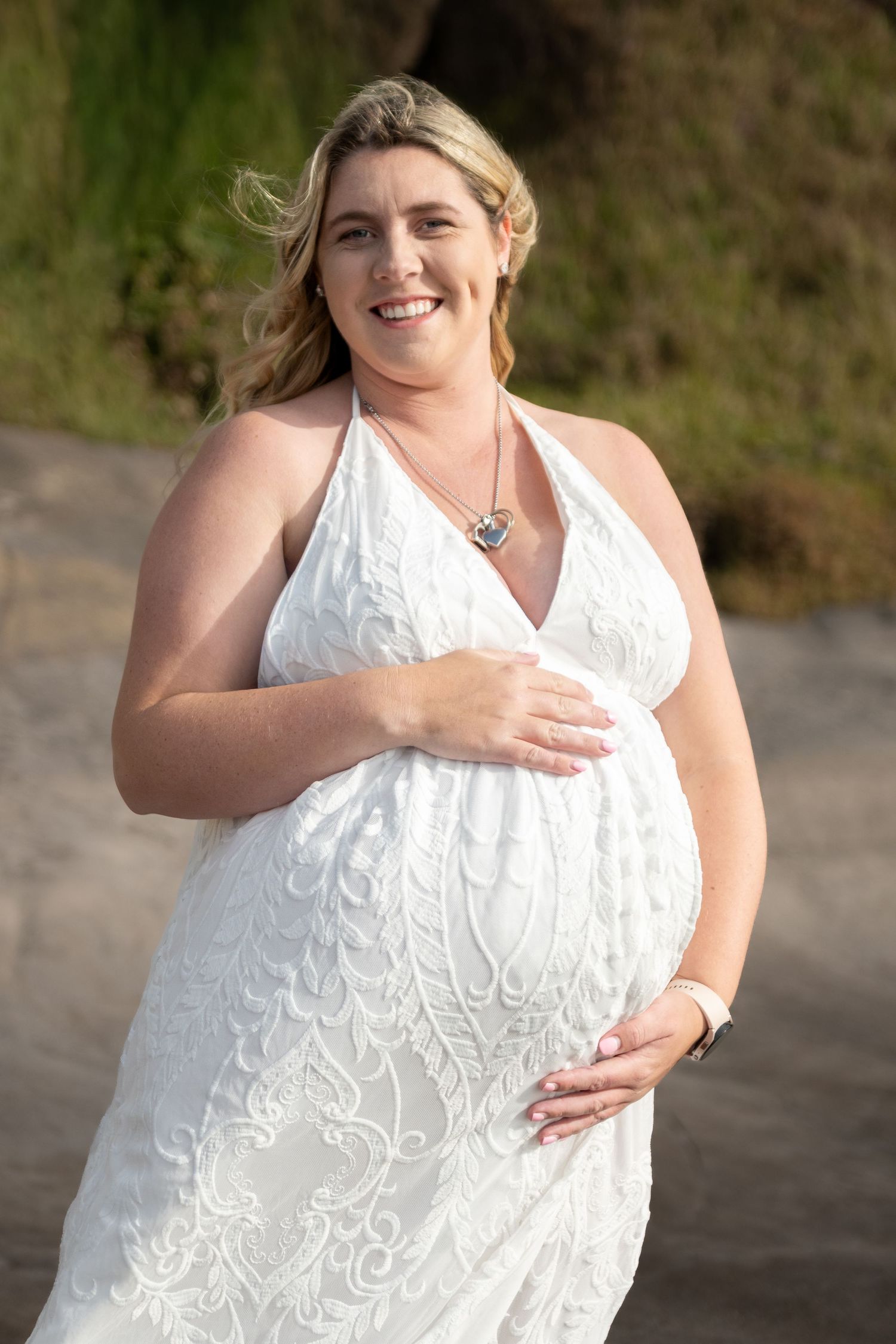 Smiling pregnant woman in white lace halter dress cradling baby bump outdoors near greenery.