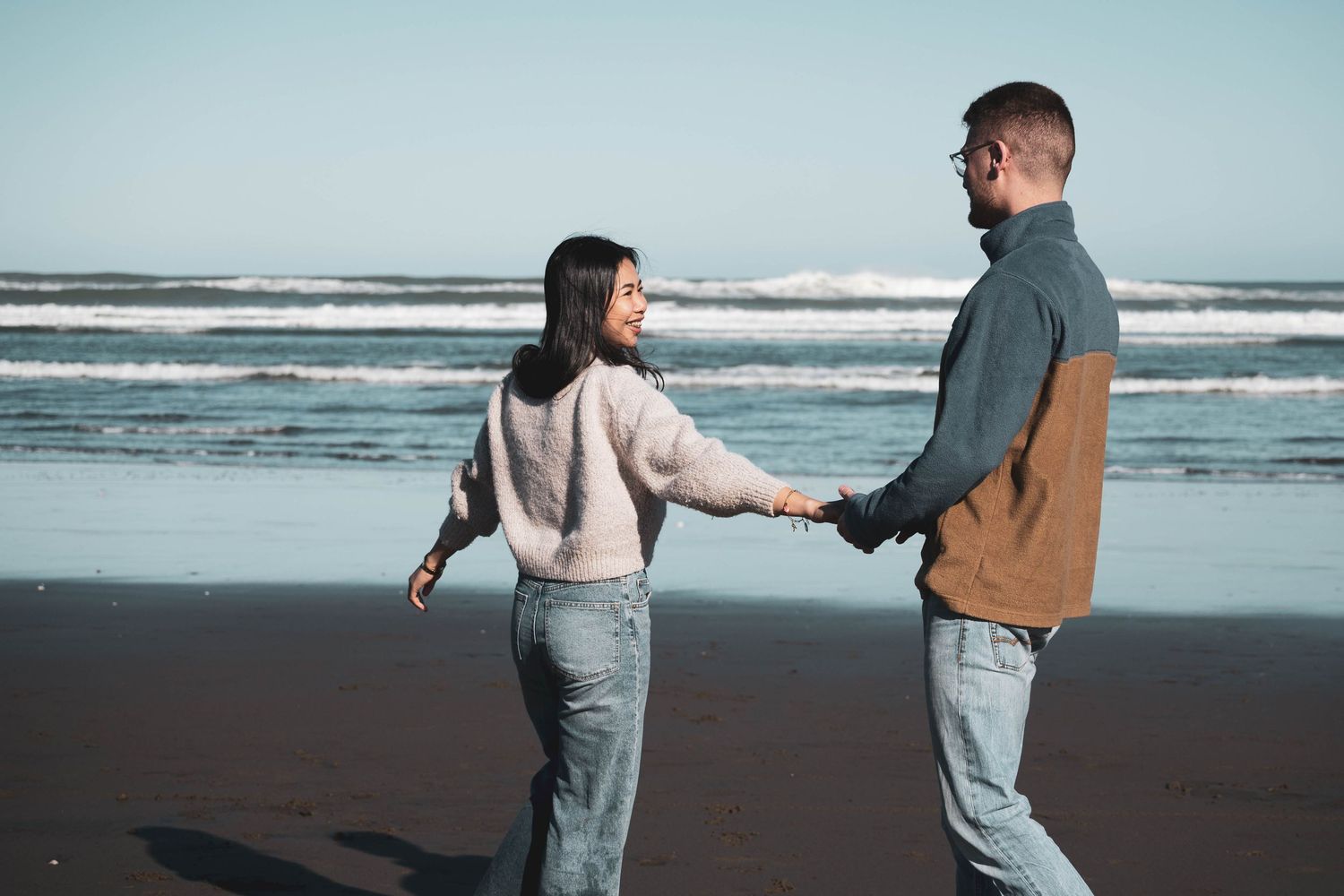Couple holding hands and walking on a black sand beach with ocean waves in the background.
