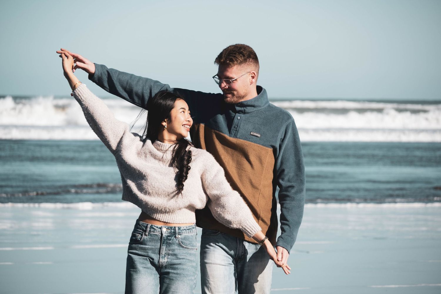A happy couple dances together on a beach, wearing casual sweaters and jeans with ocean waves in the background.