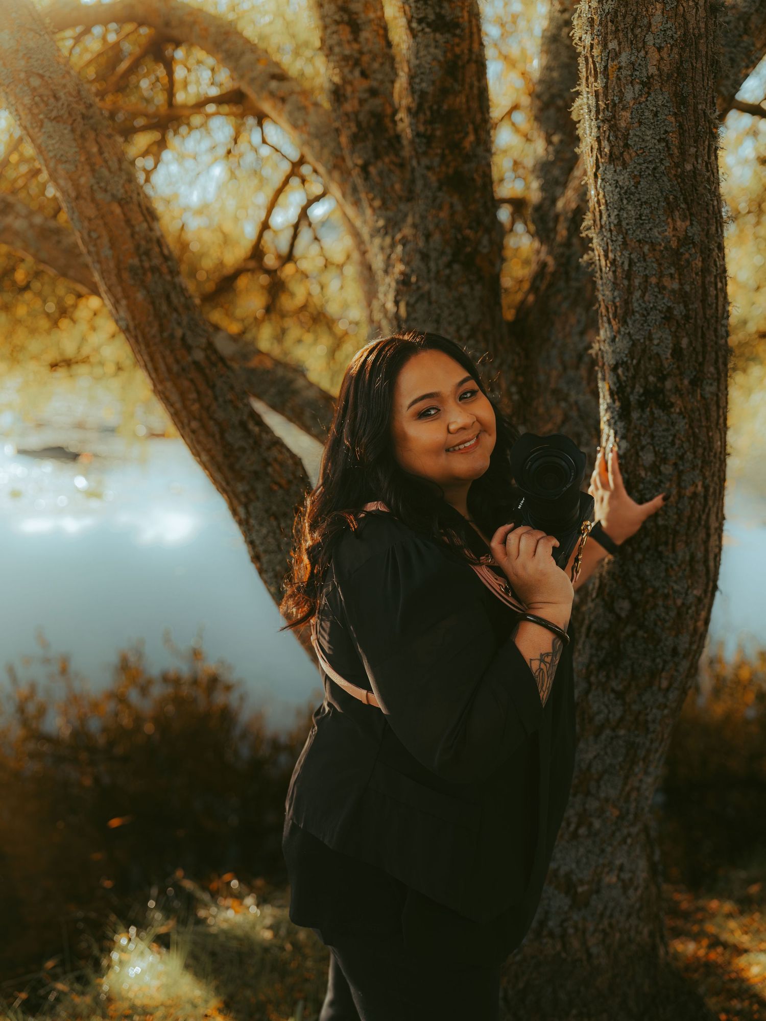 Woman in black jacket leaning against a tree by a lake, smiling warmly in a golden autumn outdoor setting.