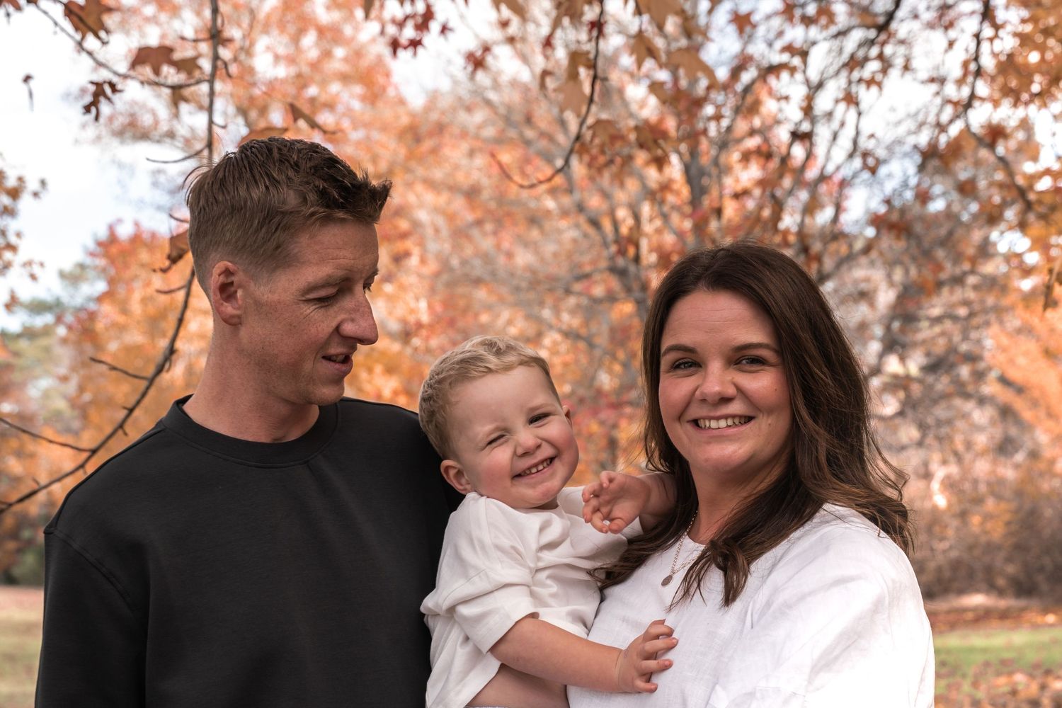 A happy young family with a smiling baby poses outdoors among vibrant autumn foliage trees.