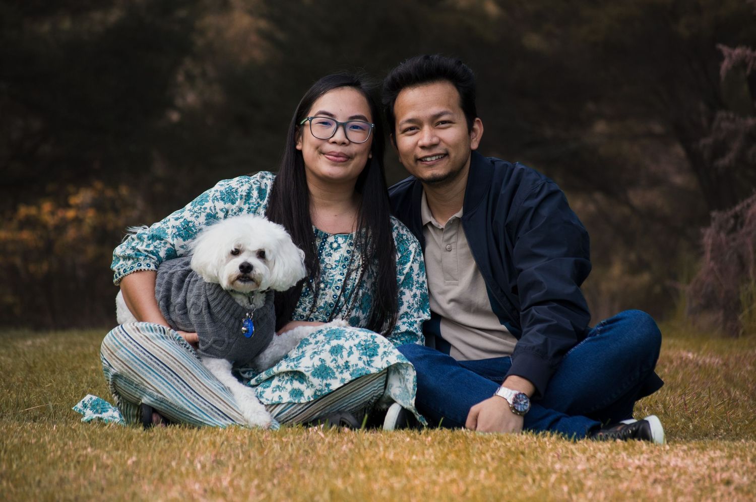 A couple sits on grass with a white fluffy dog, smiling outdoors in a park setting.