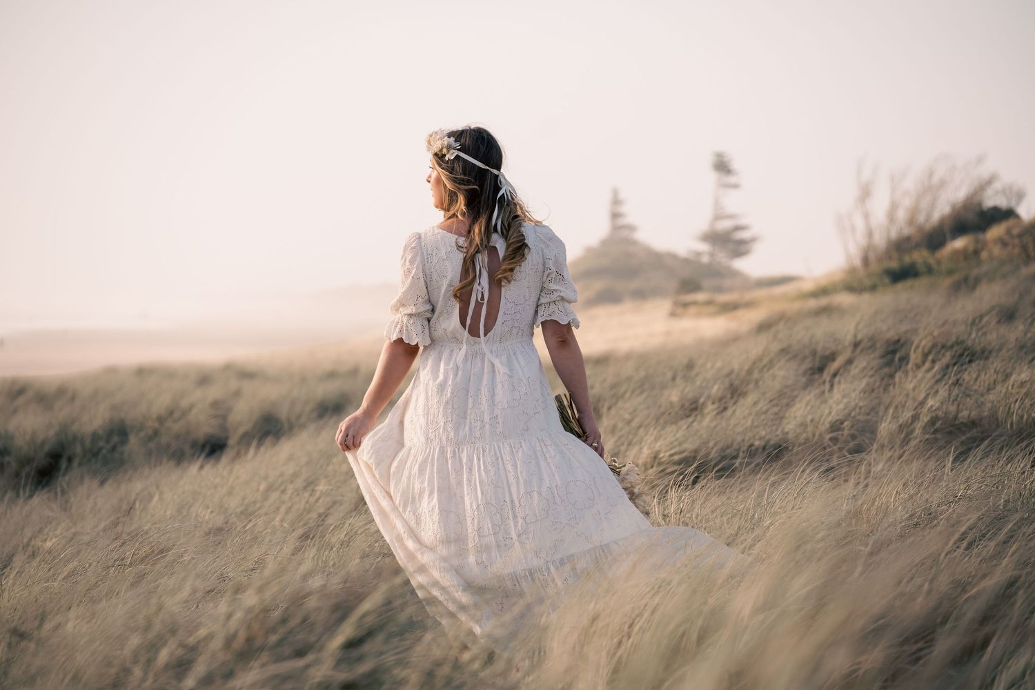 Woman in white boho dress walking through coastal dunes, hair adorned with floral crown, dress flowing in breeze.