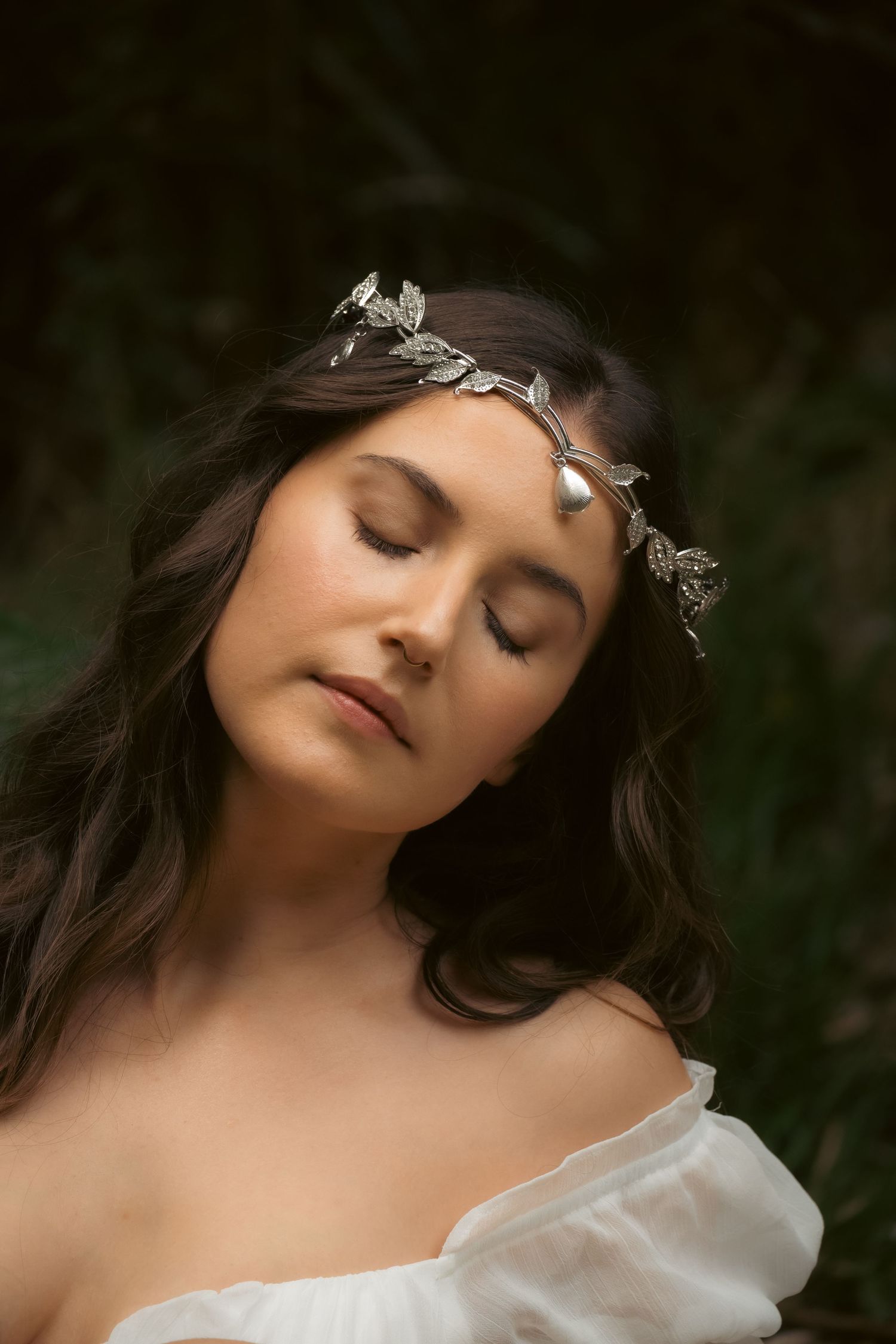 Woman with closed eyes wearing a silver leaf crown headpiece and off-shoulder white dress in a forest setting.