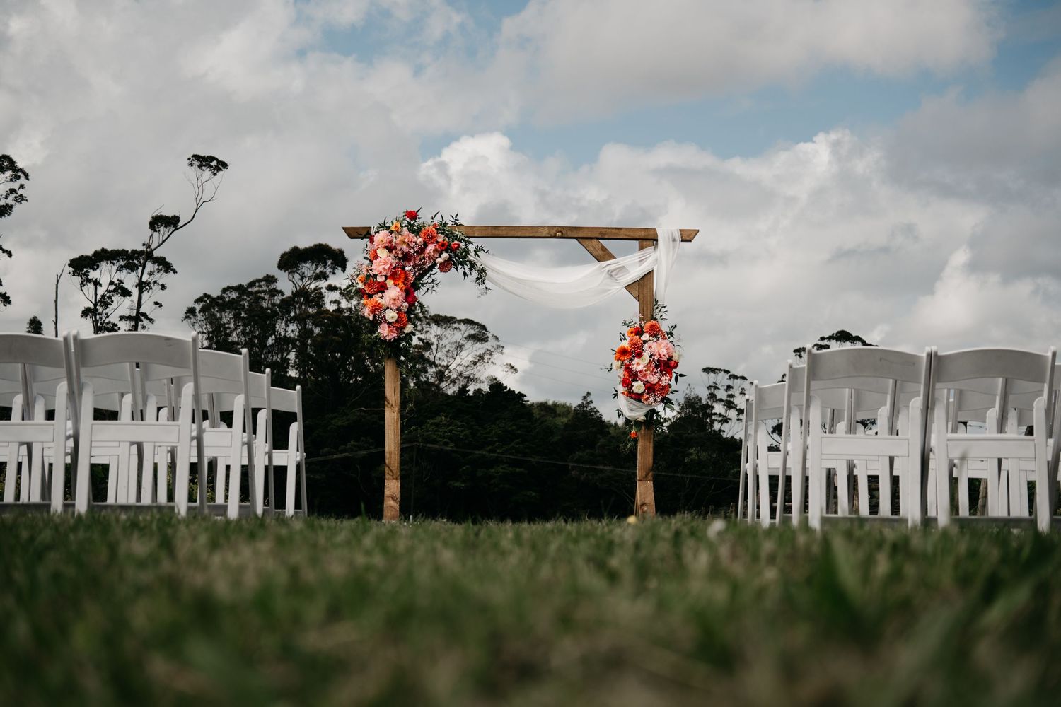Outdoor wedding ceremony setup with wooden arch adorned with pink flowers and white chairs on green lawn.