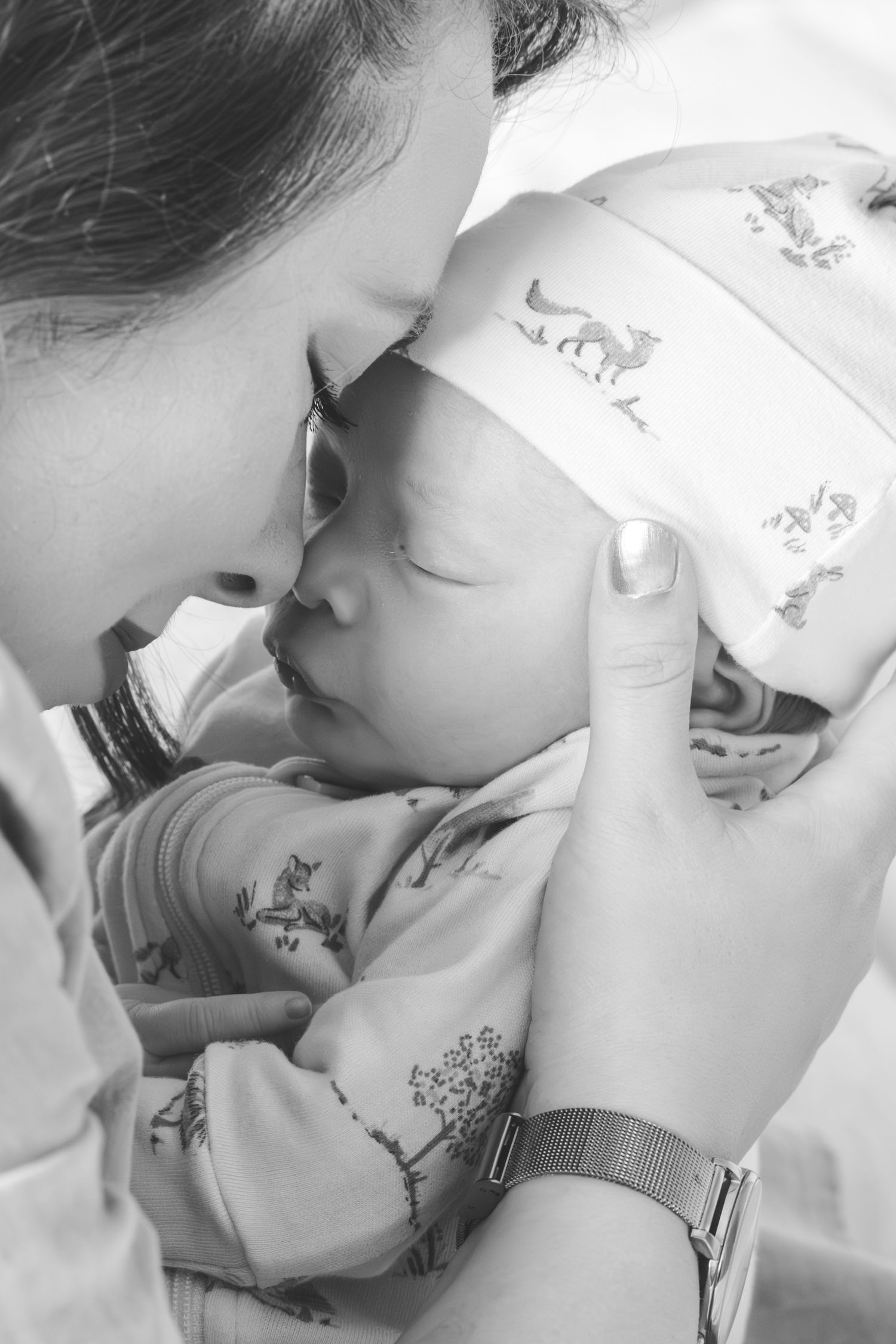 Black and white photo of a mother tenderly touching foreheads with her sleeping newborn baby wrapped in a patterned blanket.