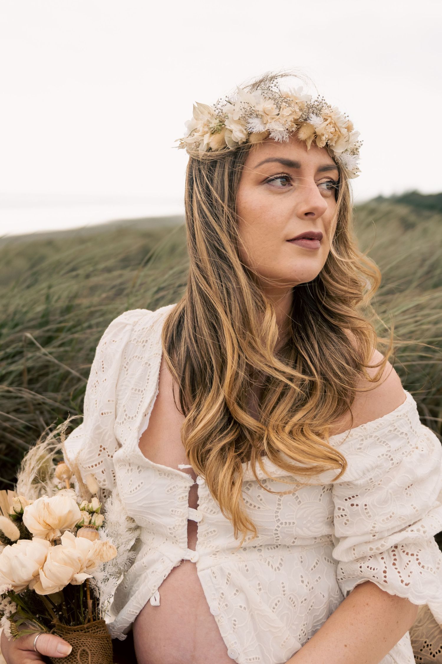 Pregnant woman in white off-shoulder dress and floral crown holds bouquet outdoors near grassy dunes.