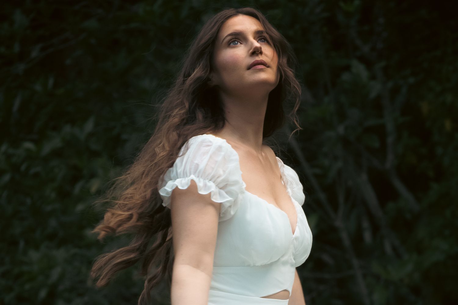 Young woman with long brown hair in white ruffled crop top poses gracefully outdoors against lush green foliage.