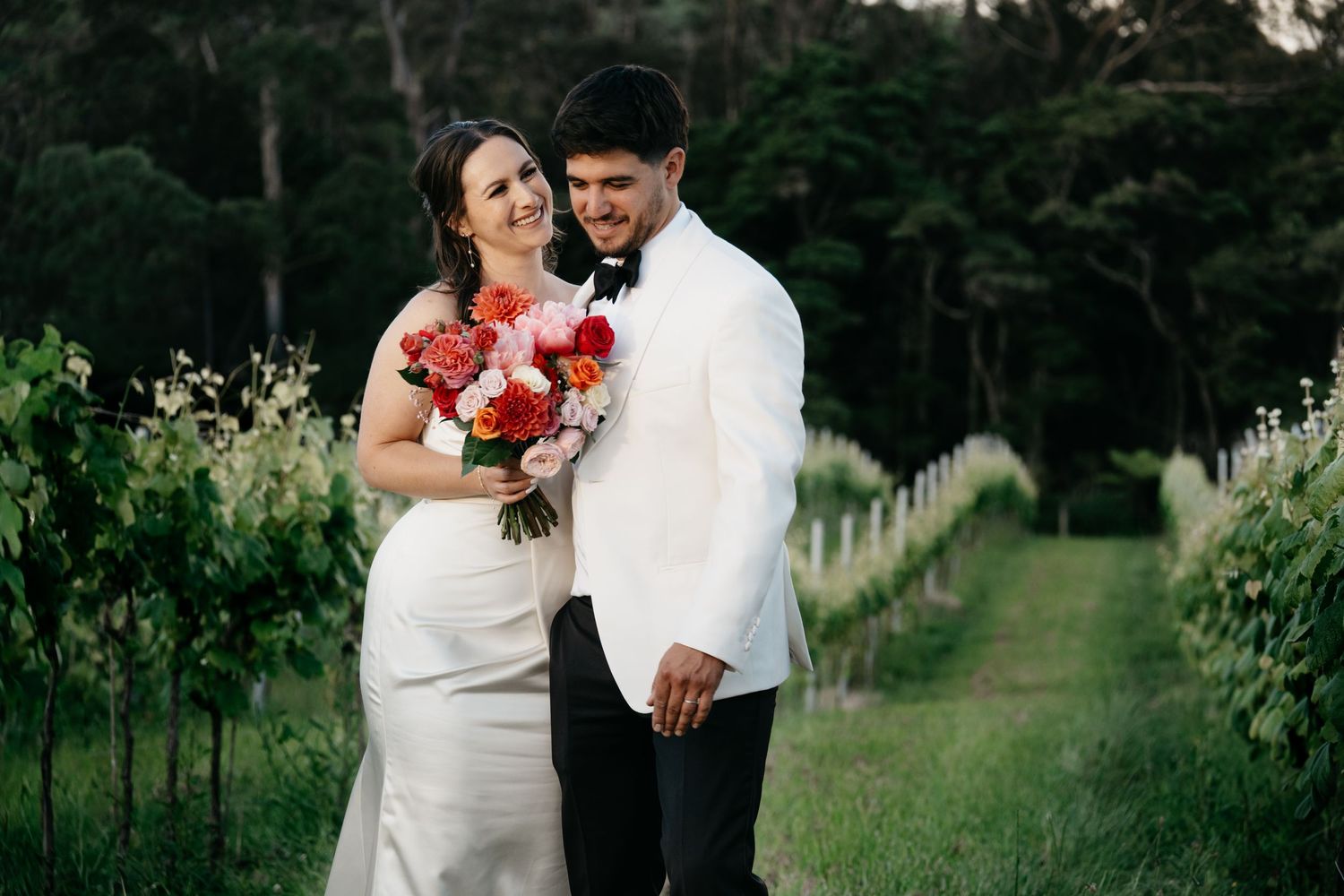 Bride and groom pose in vineyard, she holds red bouquet in white gown, he wears white tuxedo jacket.