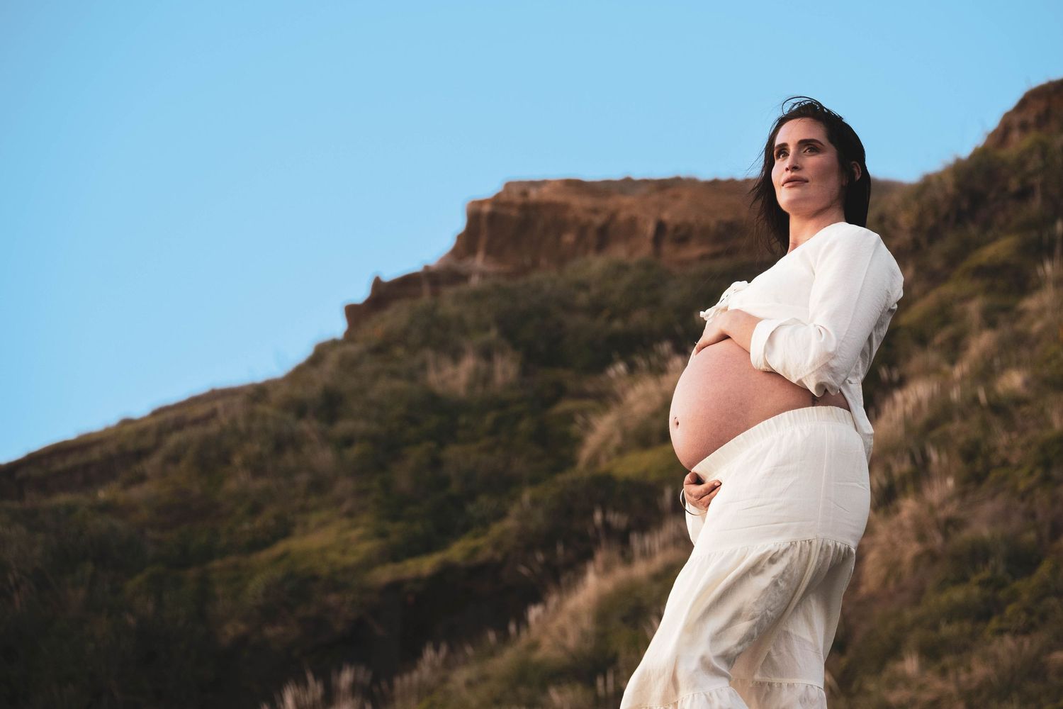 Pregnant woman in white dress cradling baby bump outdoors against rocky hillside under blue sky.