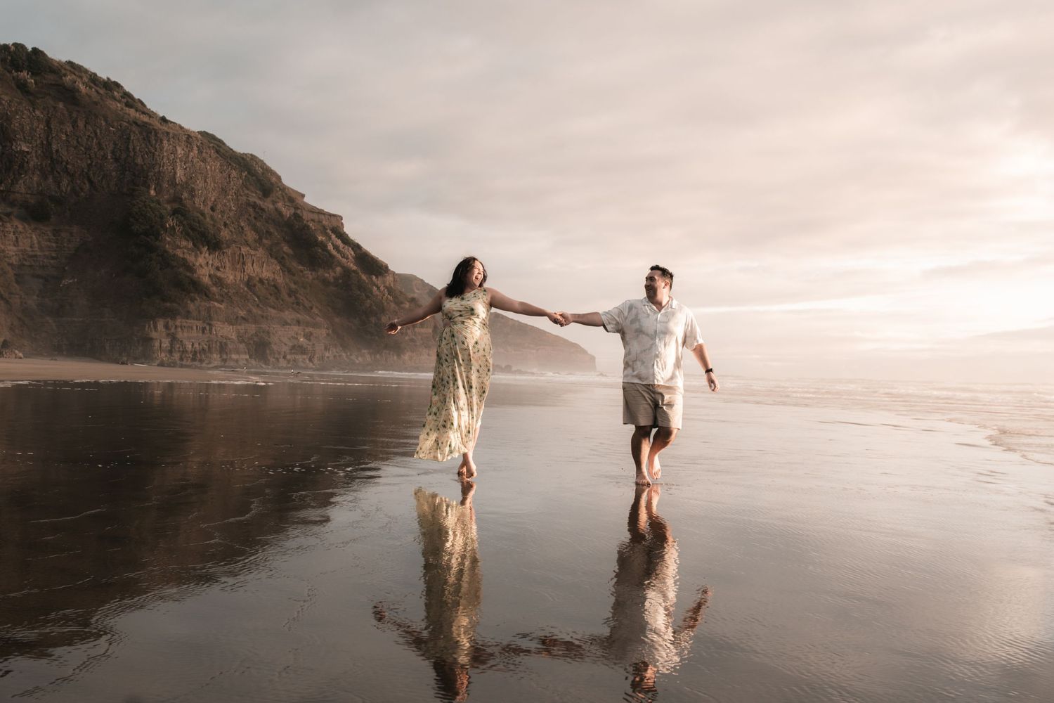 Couple holding hands walking on reflective wet beach at sunset with dramatic cliffs in background.