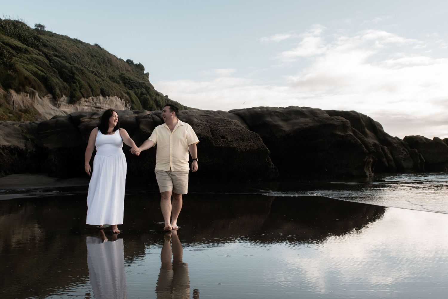 Couple holding hands walking on a reflective beach with rocky cliffs at sunset.