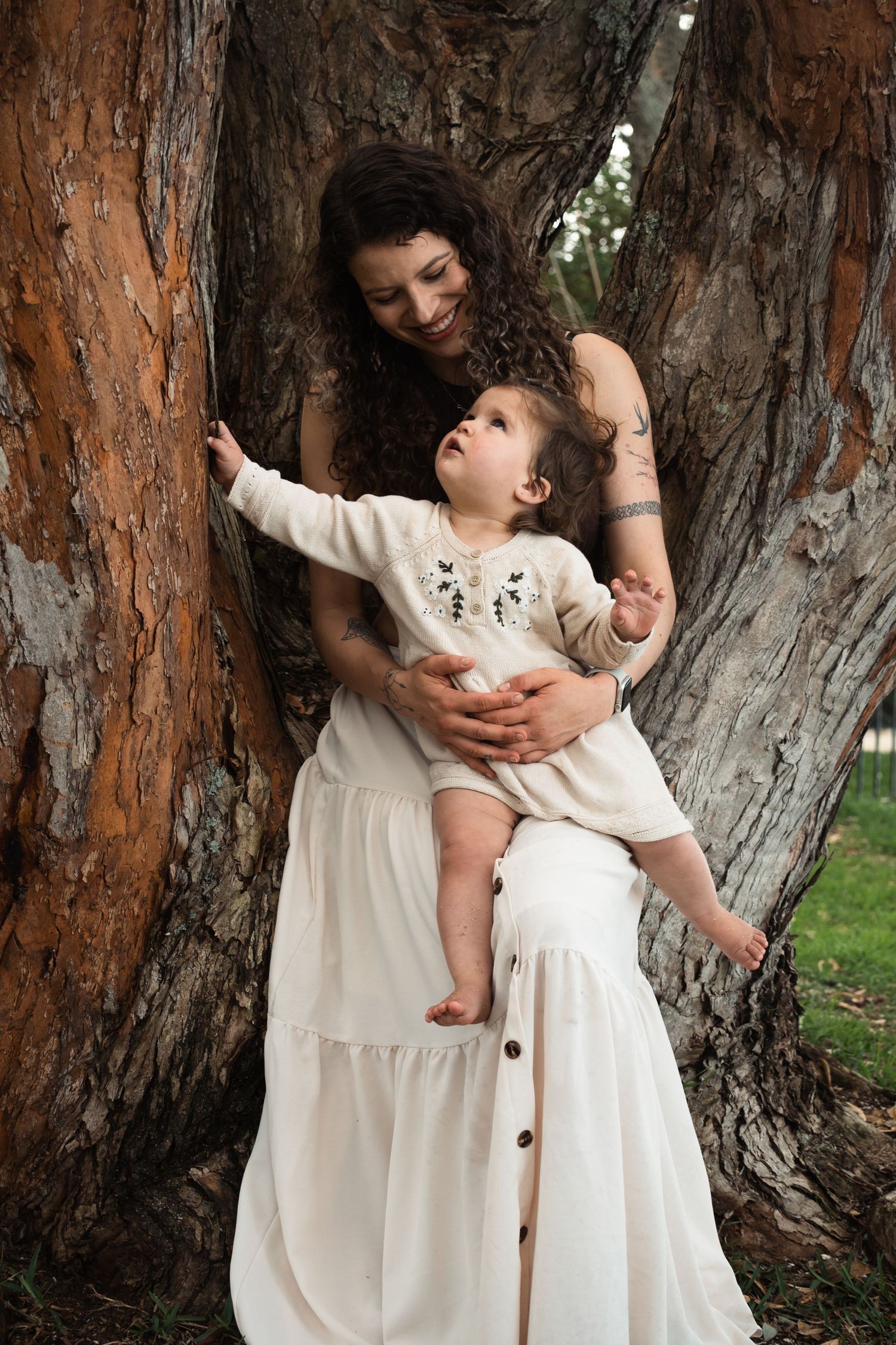 A curly-haired mother in a white dress holds her baby near a large tree trunk outdoors.