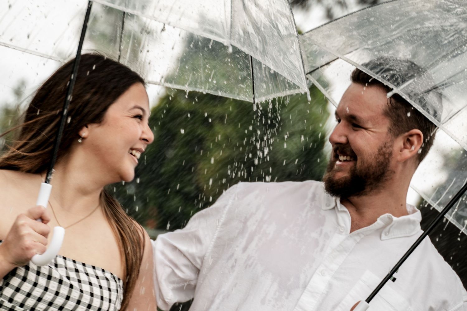 Happy couple laughing together under clear umbrellas in the rain, sharing a joyful romantic moment outdoors.