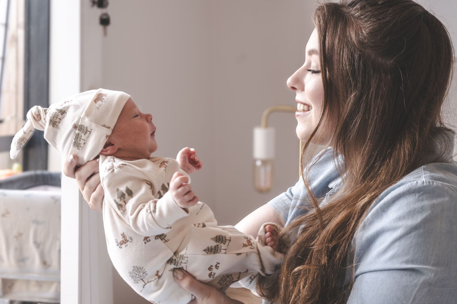 A smiling mother holds her newborn baby dressed in a floral outfit and hat near a bright window.