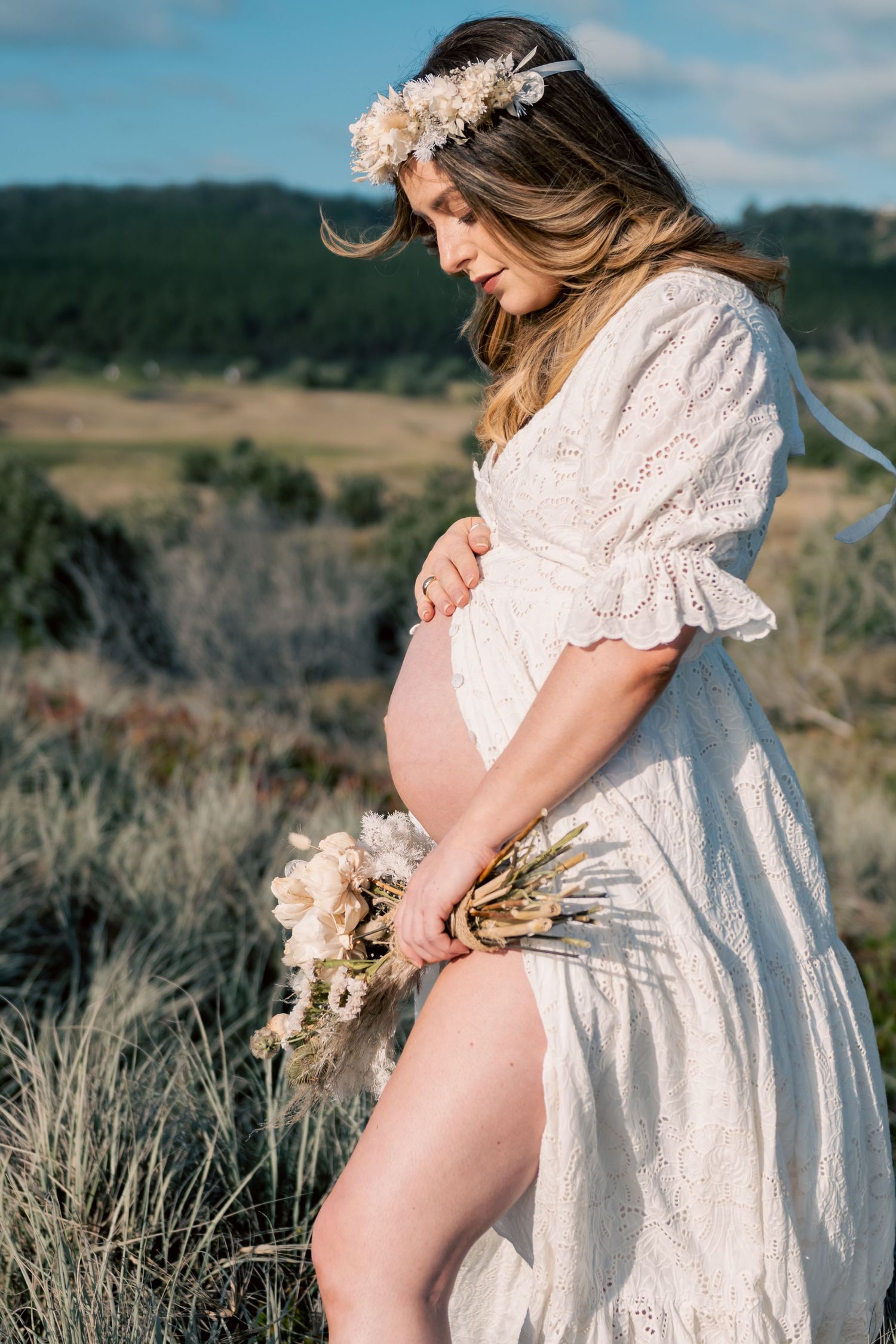Pregnant woman in white lace dress and floral crown holds bouquet outdoors in scenic field.