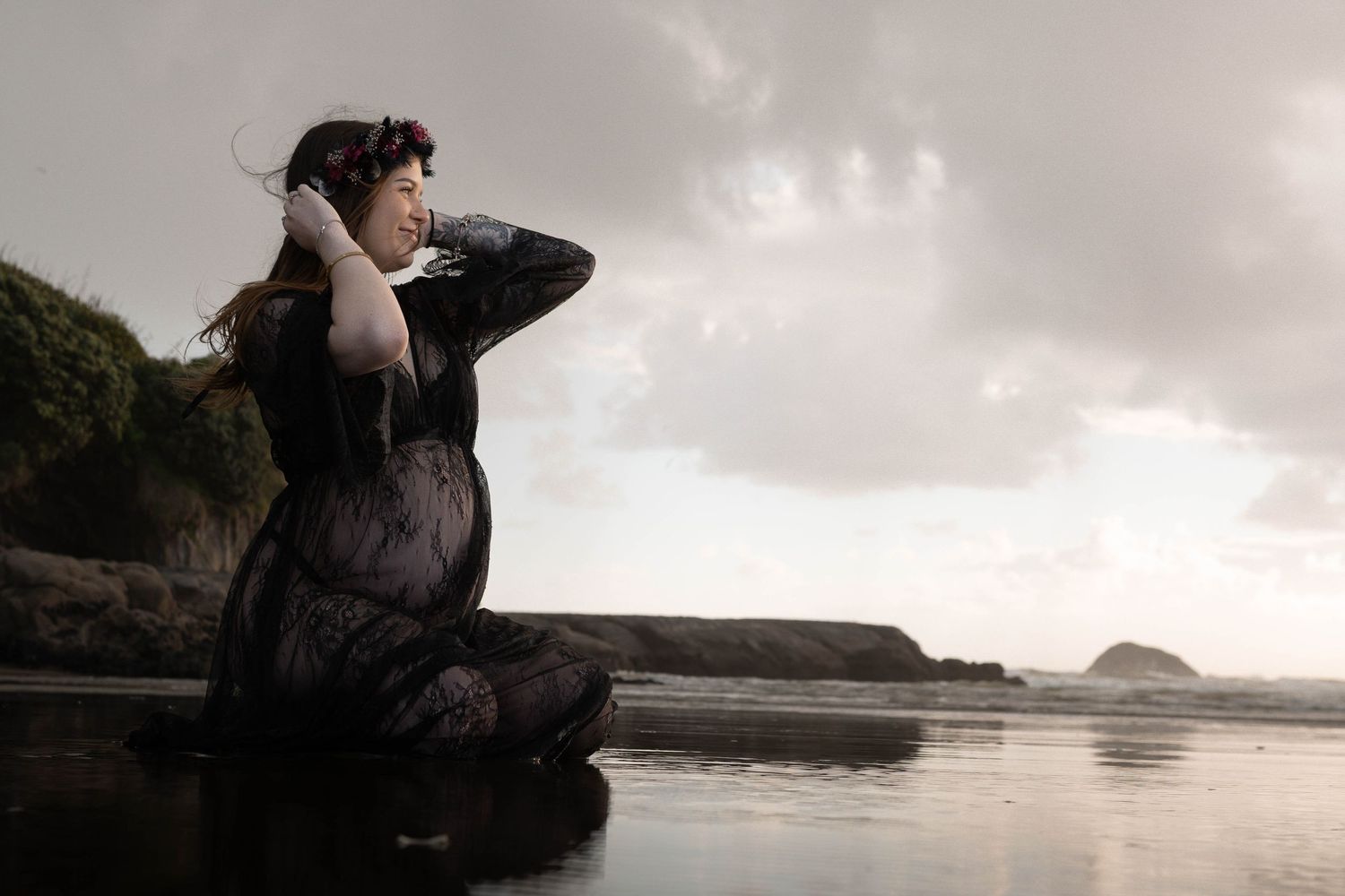 Pregnant woman in black lace dress poses gracefully on a reflective beach at dusk with cloudy skies.