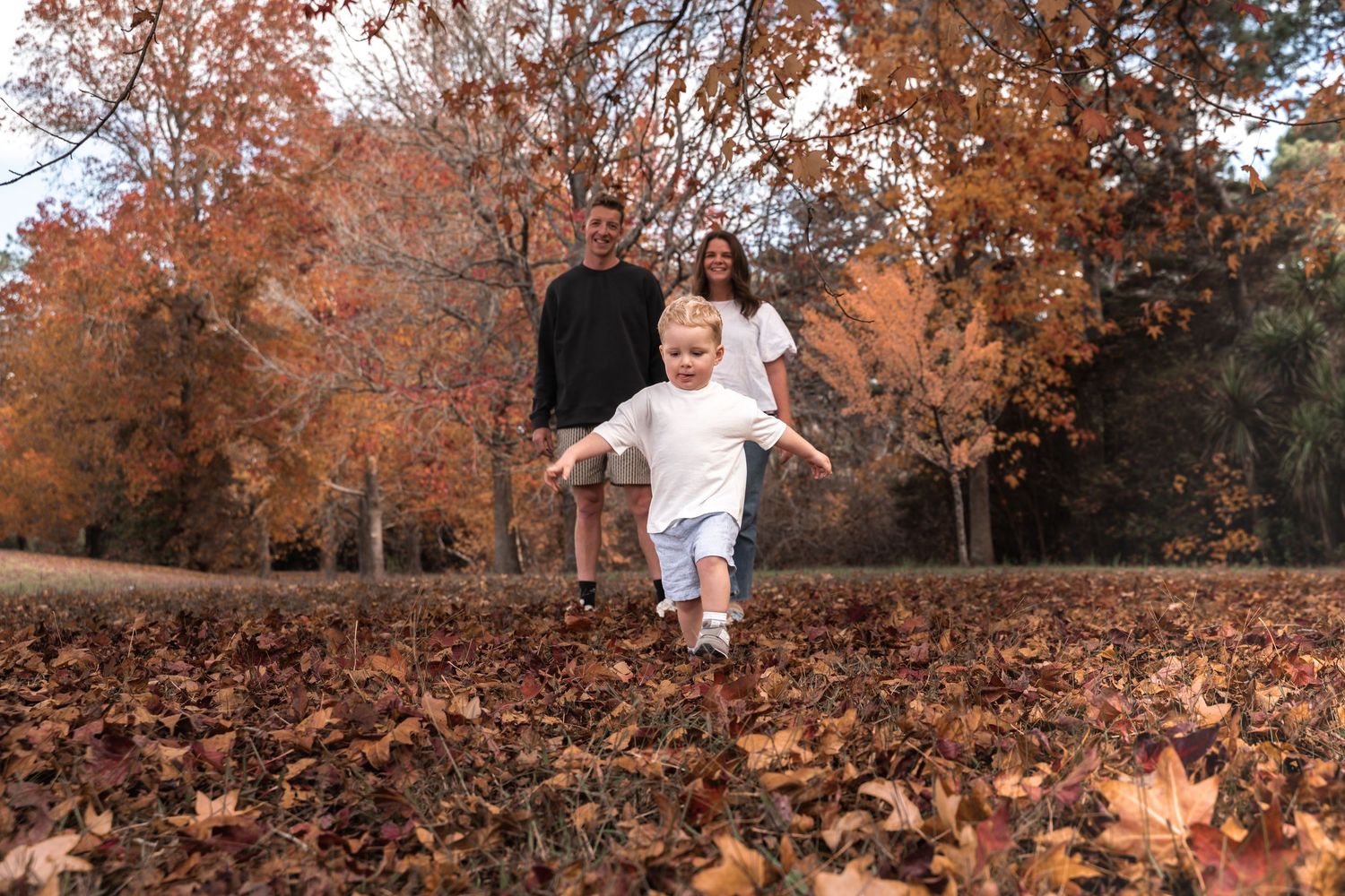 A joyful family plays in autumn leaves in a park, with a young boy running toward the camera.