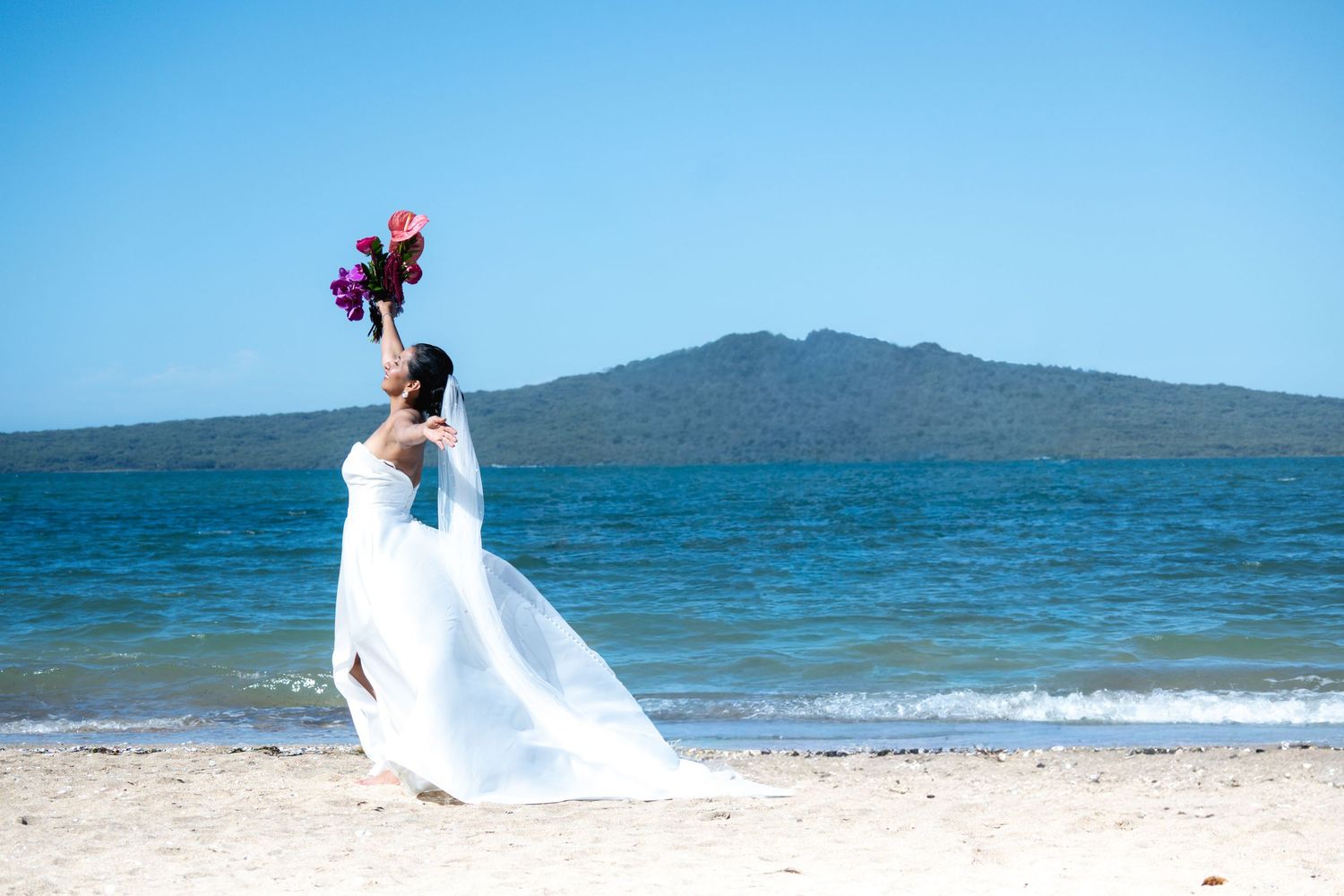 Bride in white gown tosses purple bouquet on sandy beach with blue ocean and volcanic island backdrop.