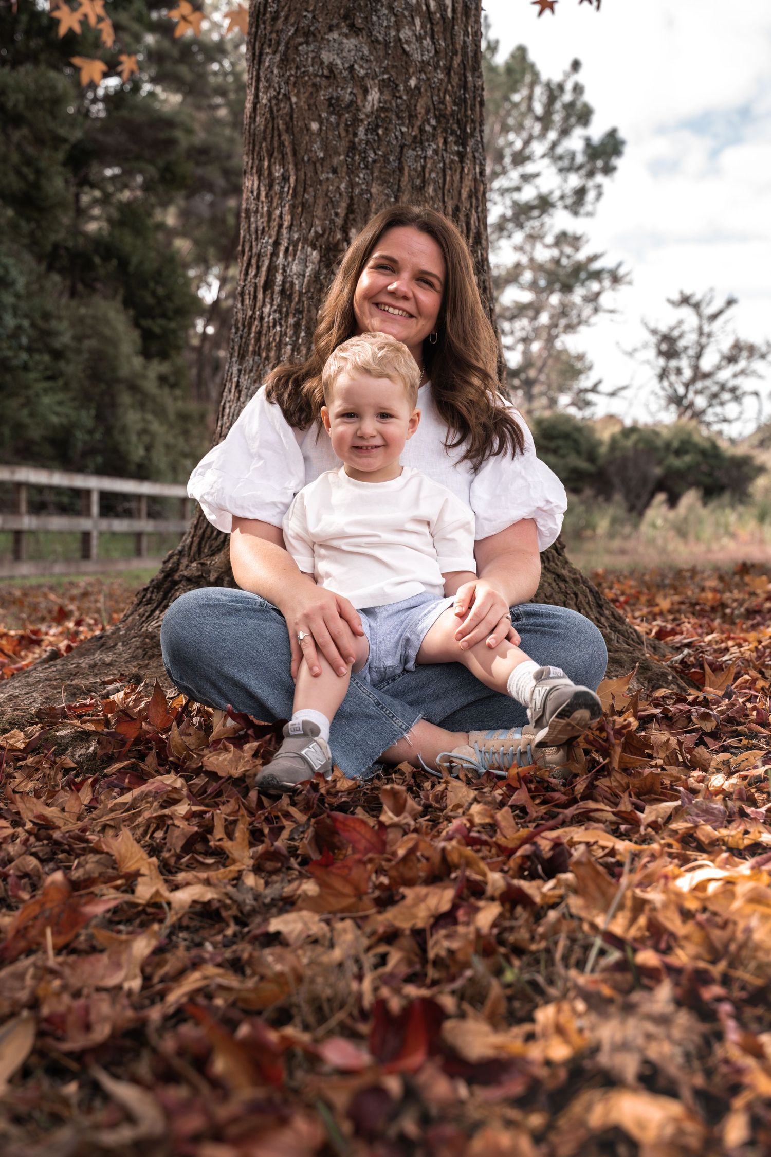 A smiling mother and toddler sit together against a tree trunk surrounded by autumn leaves in a park.