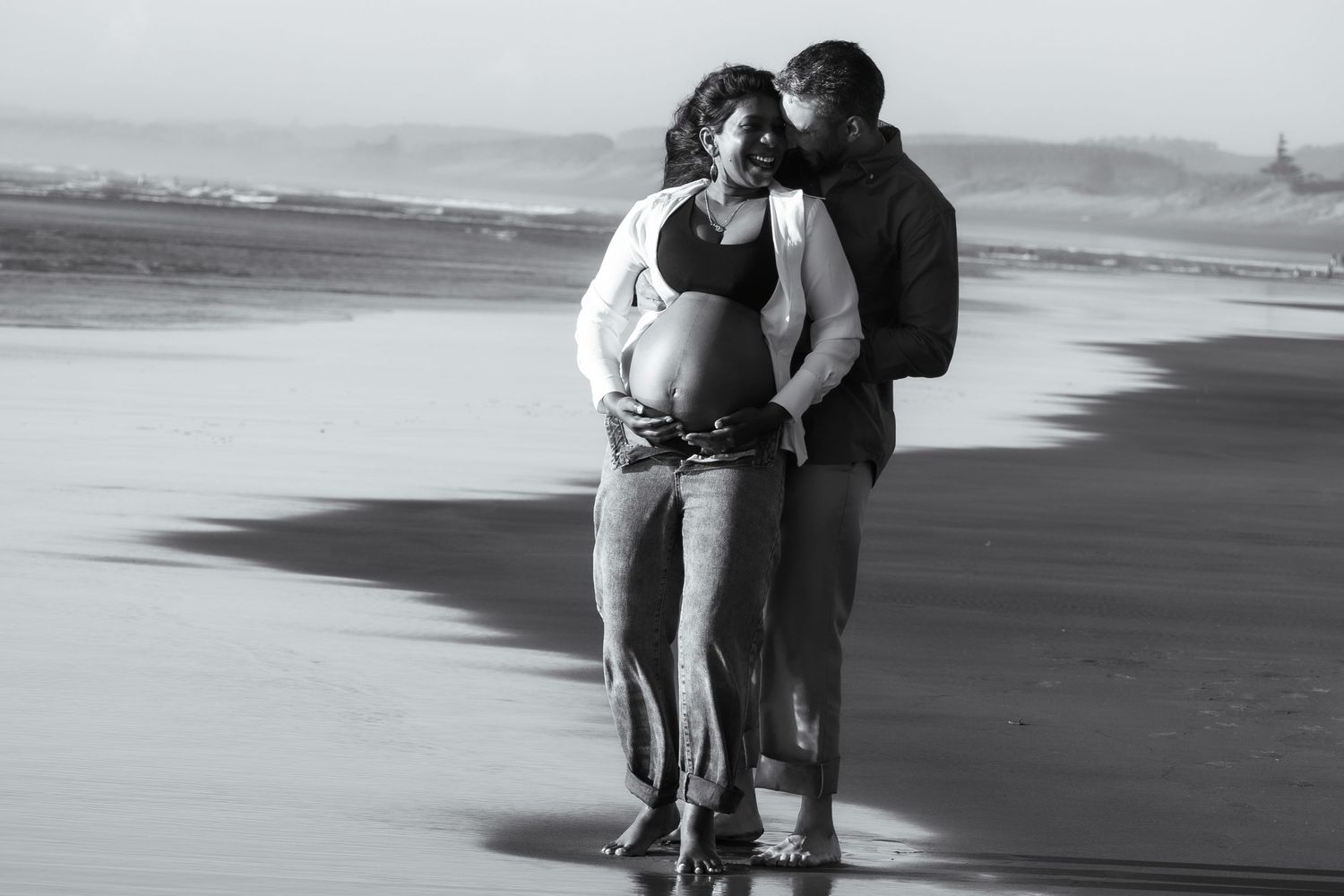 Black and white photo of a pregnant couple embracing on a beach, the man kissing her head as she holds her baby bump.