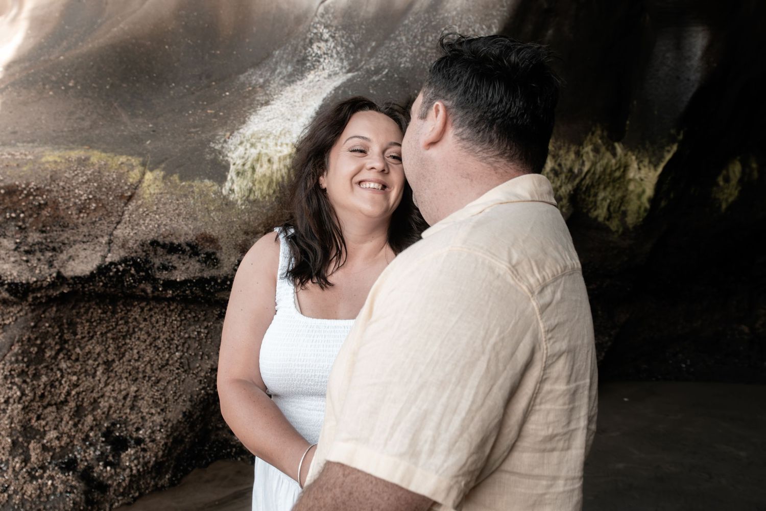 Couple smiling and laughing together on a rocky beach, woman in white dress and man in beige shirt.