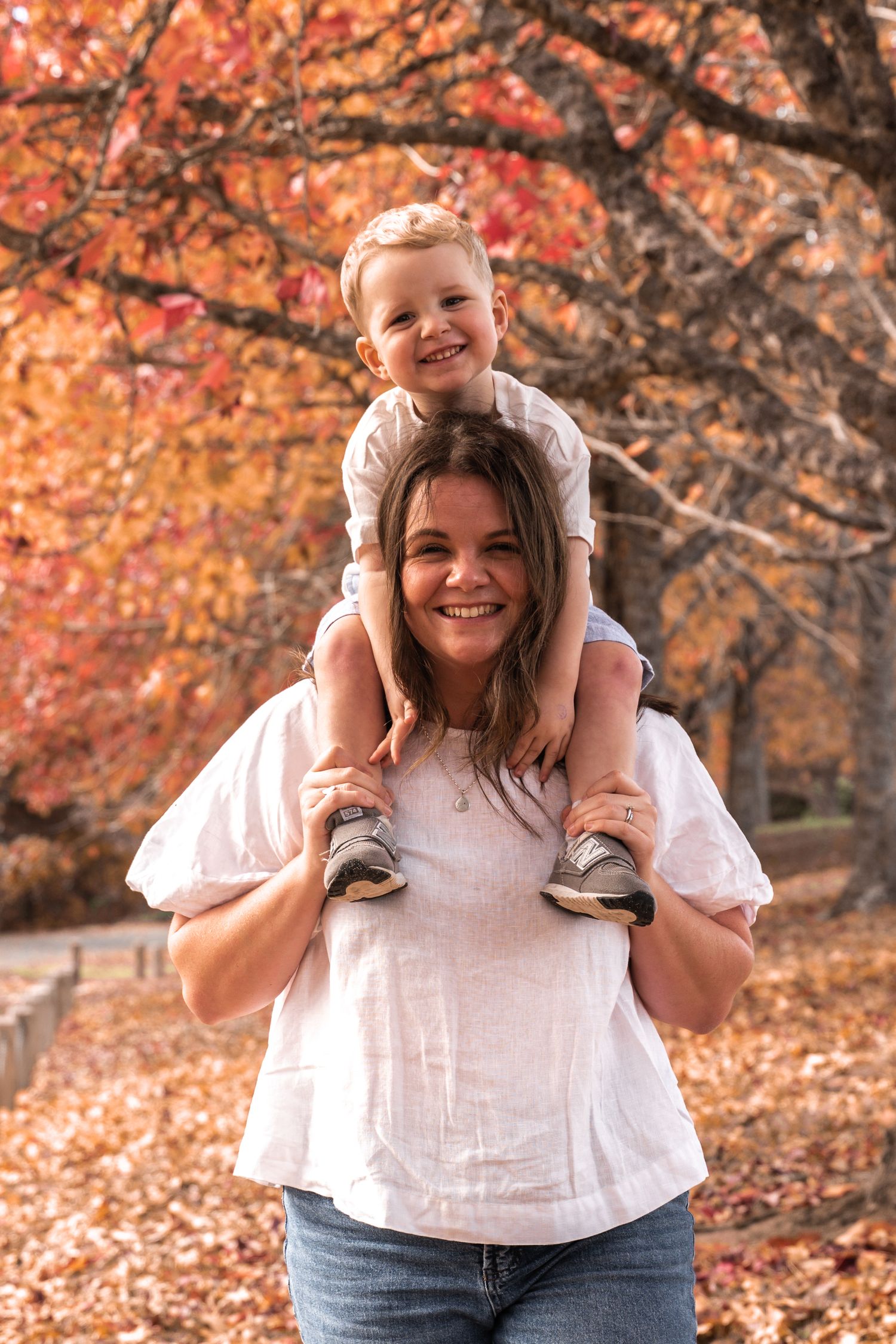 A smiling woman carries a happy young boy on her shoulders outdoors amid vibrant autumn foliage.