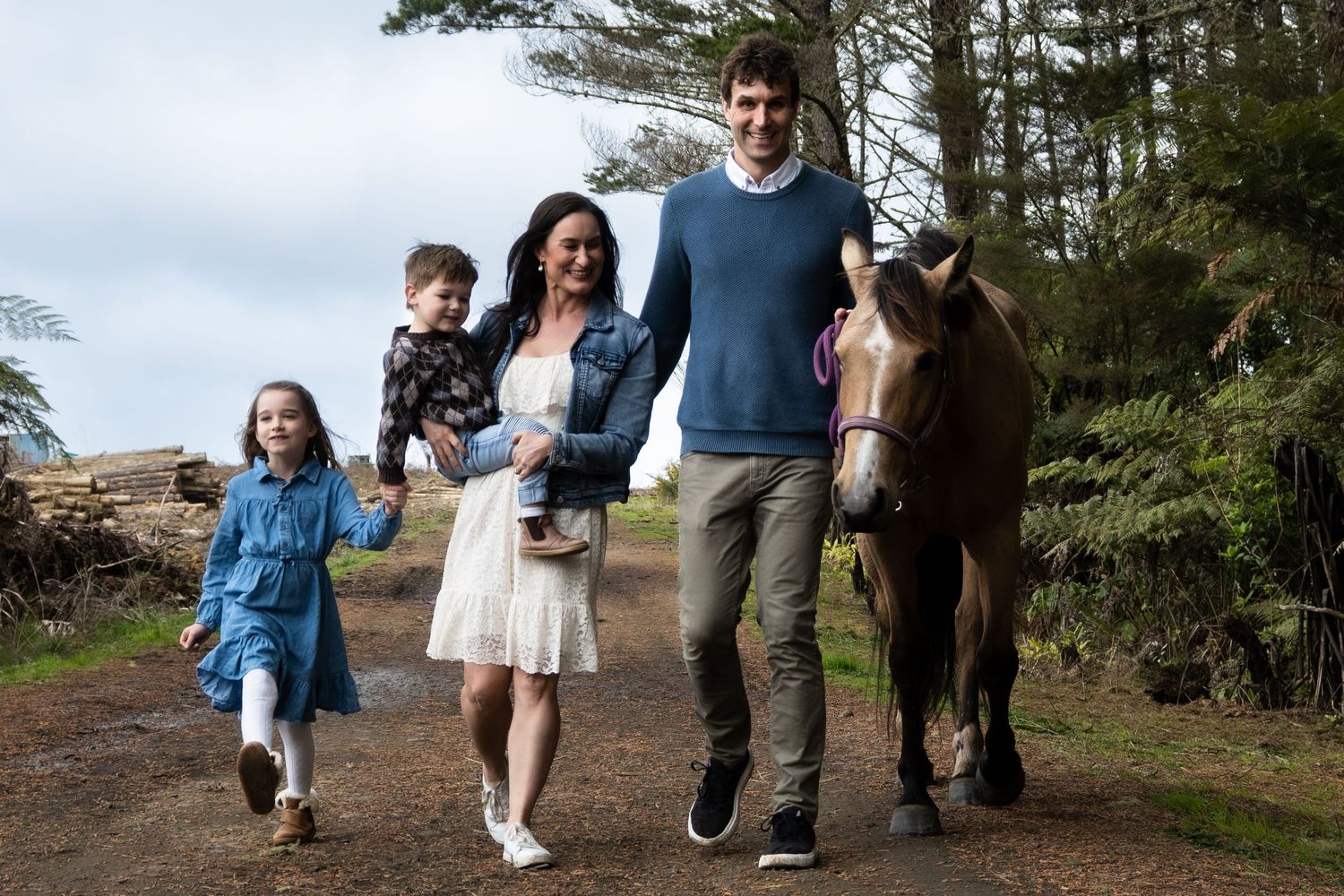 Family of four walking with a brown horse on a rural path surrounded by lush green trees.