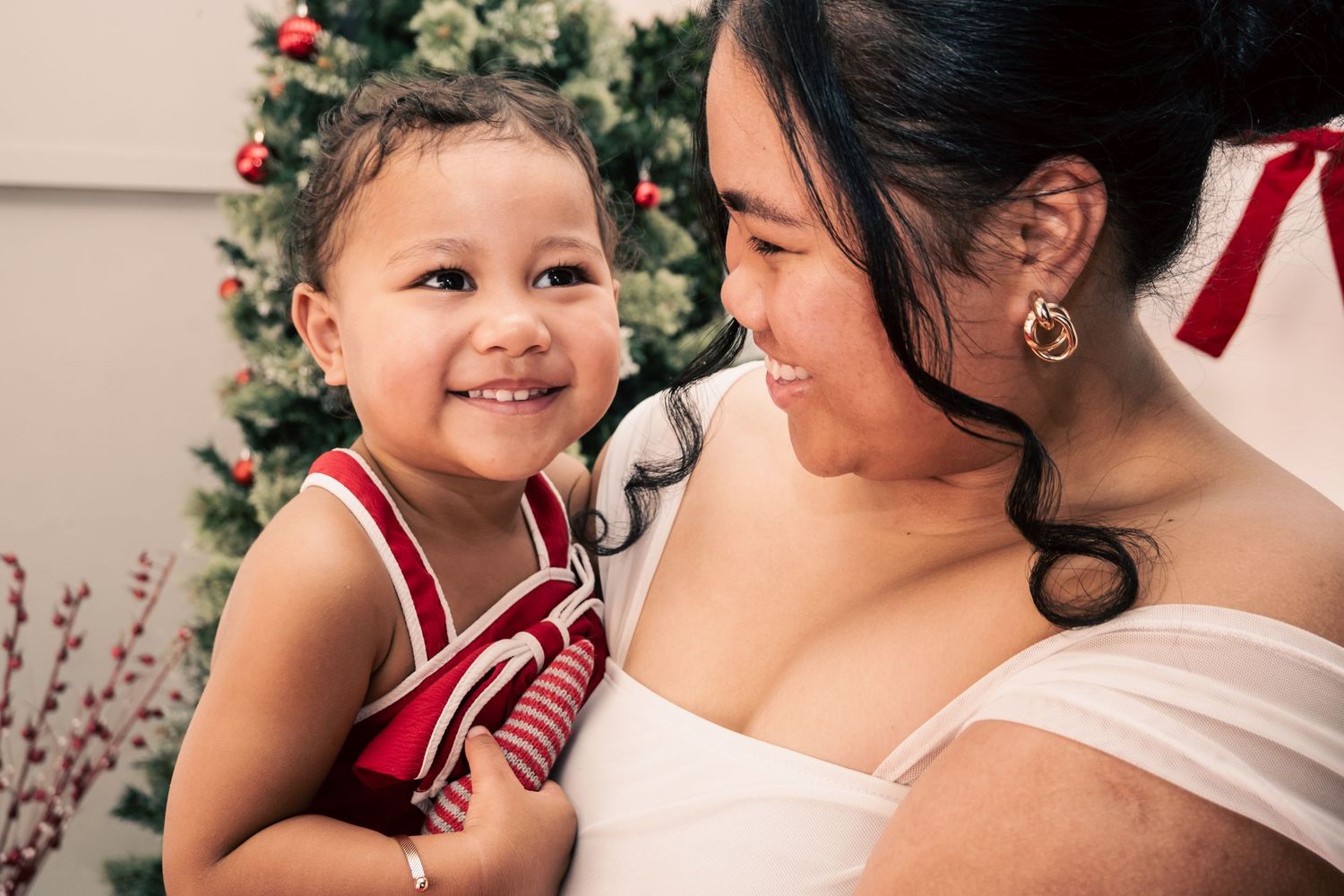 A smiling toddler in red overalls being held by a woman near a decorated Christmas tree.