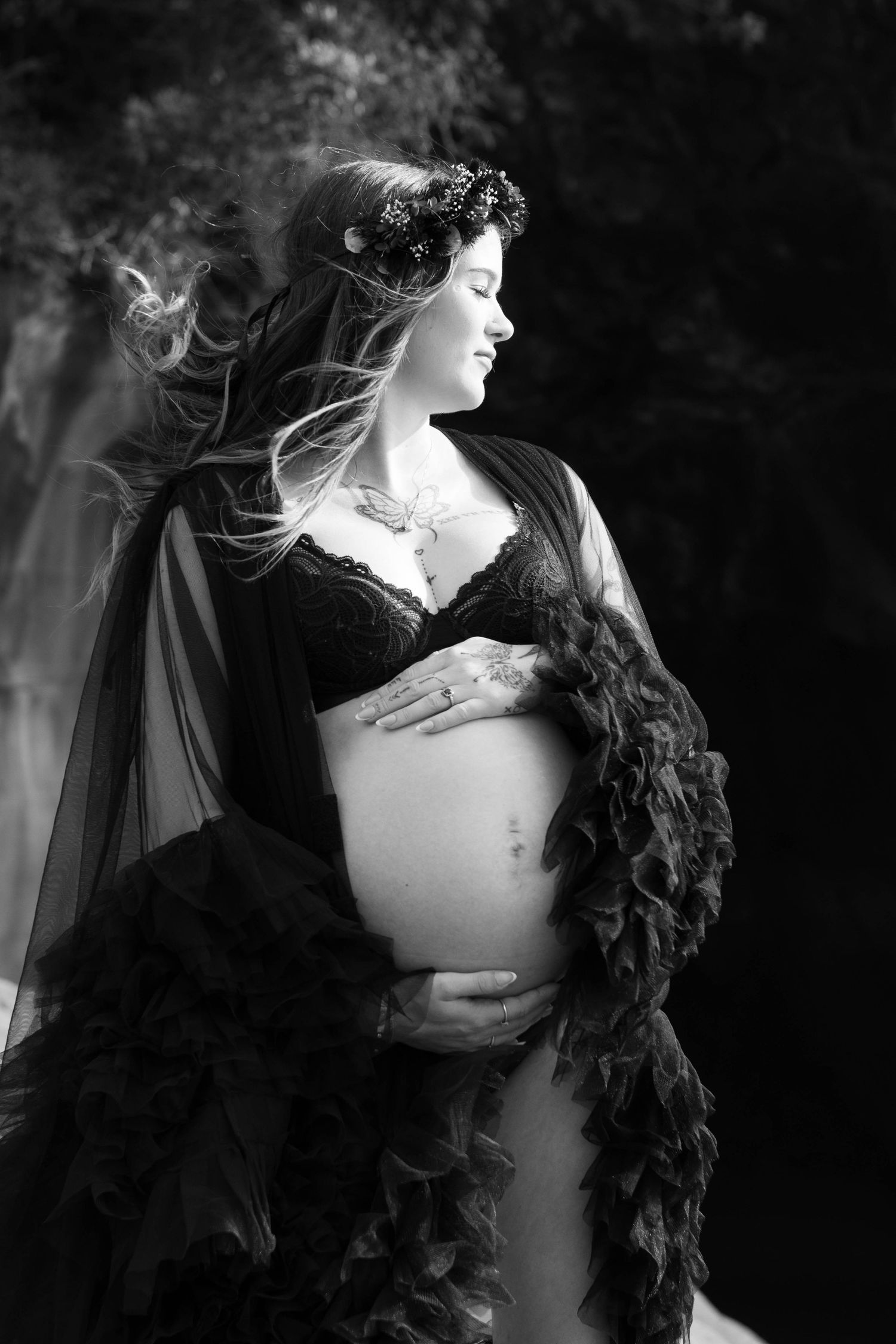 Black and white maternity photo of pregnant woman in lace bra and flowing dark robe, wearing floral crown outdoors.