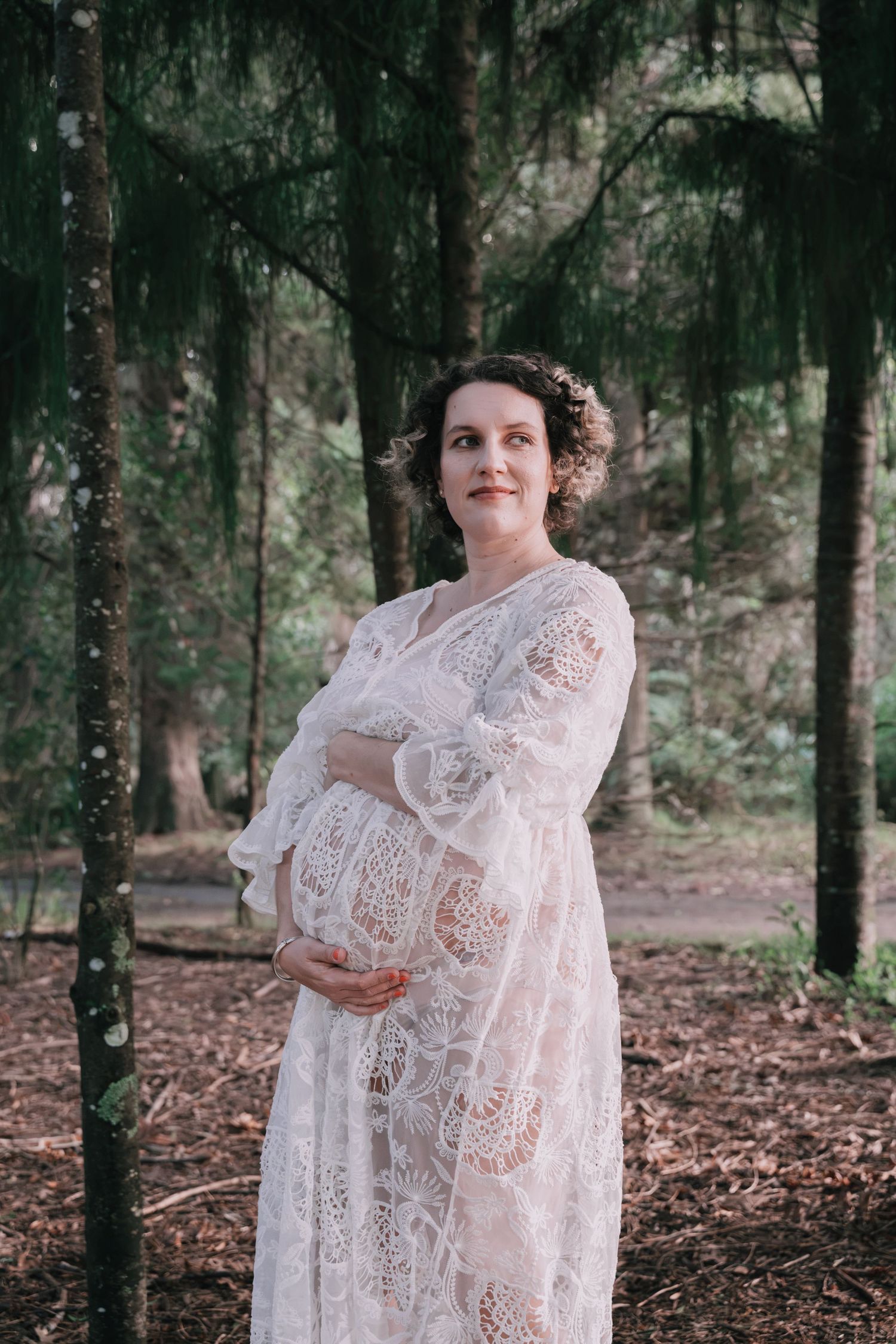 Pregnant woman in white lace dress cradling baby bump among tall trees in a serene forest maternity shoot.