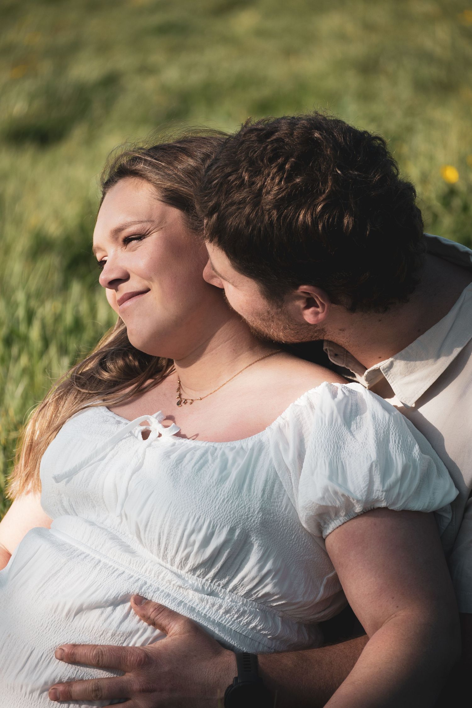A man tenderly kisses a smiling pregnant woman in a white dress outdoors in a sunny green field.