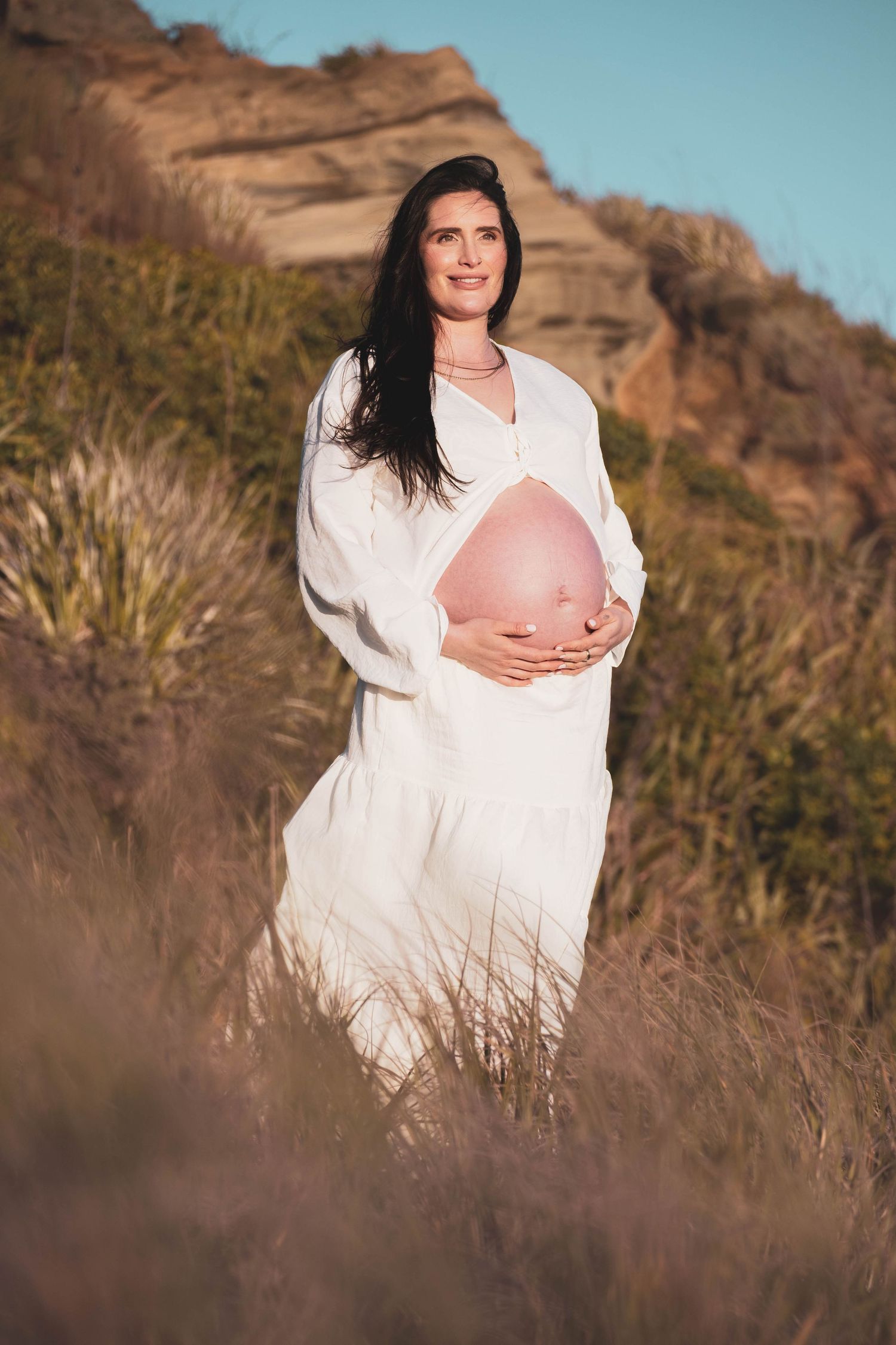 Pregnant woman in white dress cradling her baby bump outdoors in a natural landscape with rocky hills and tall grass.