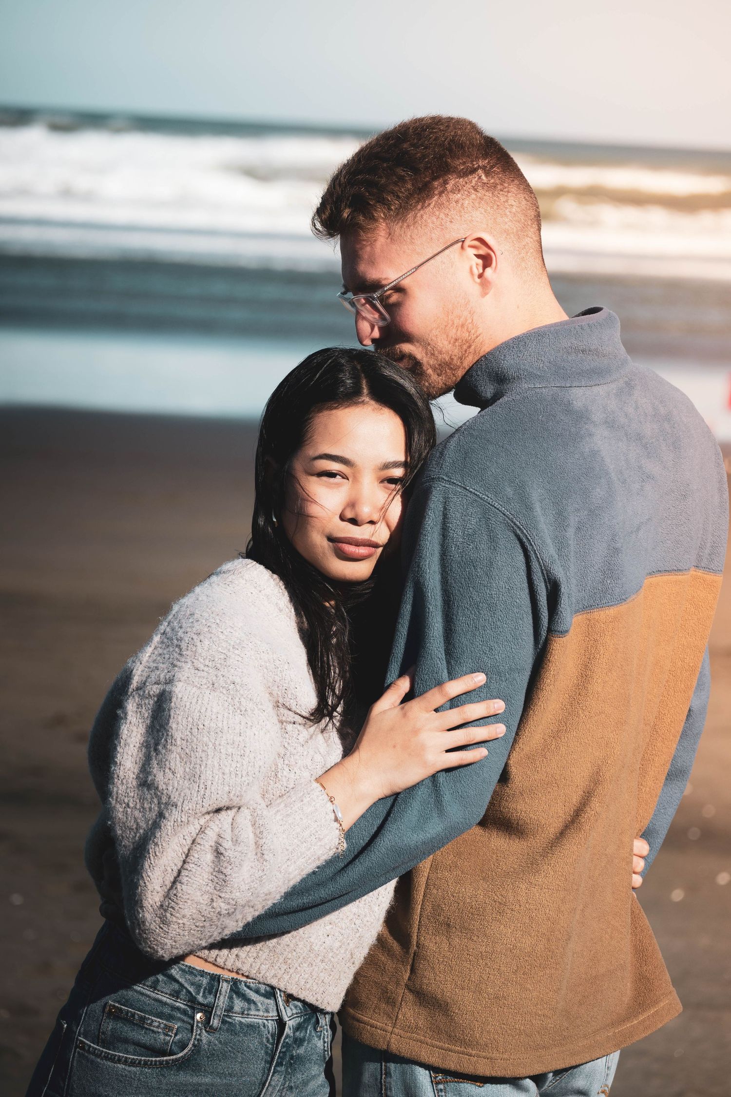 A couple embraces on a beach, the woman in a cozy sweater gazing softly while waves crash in the background.