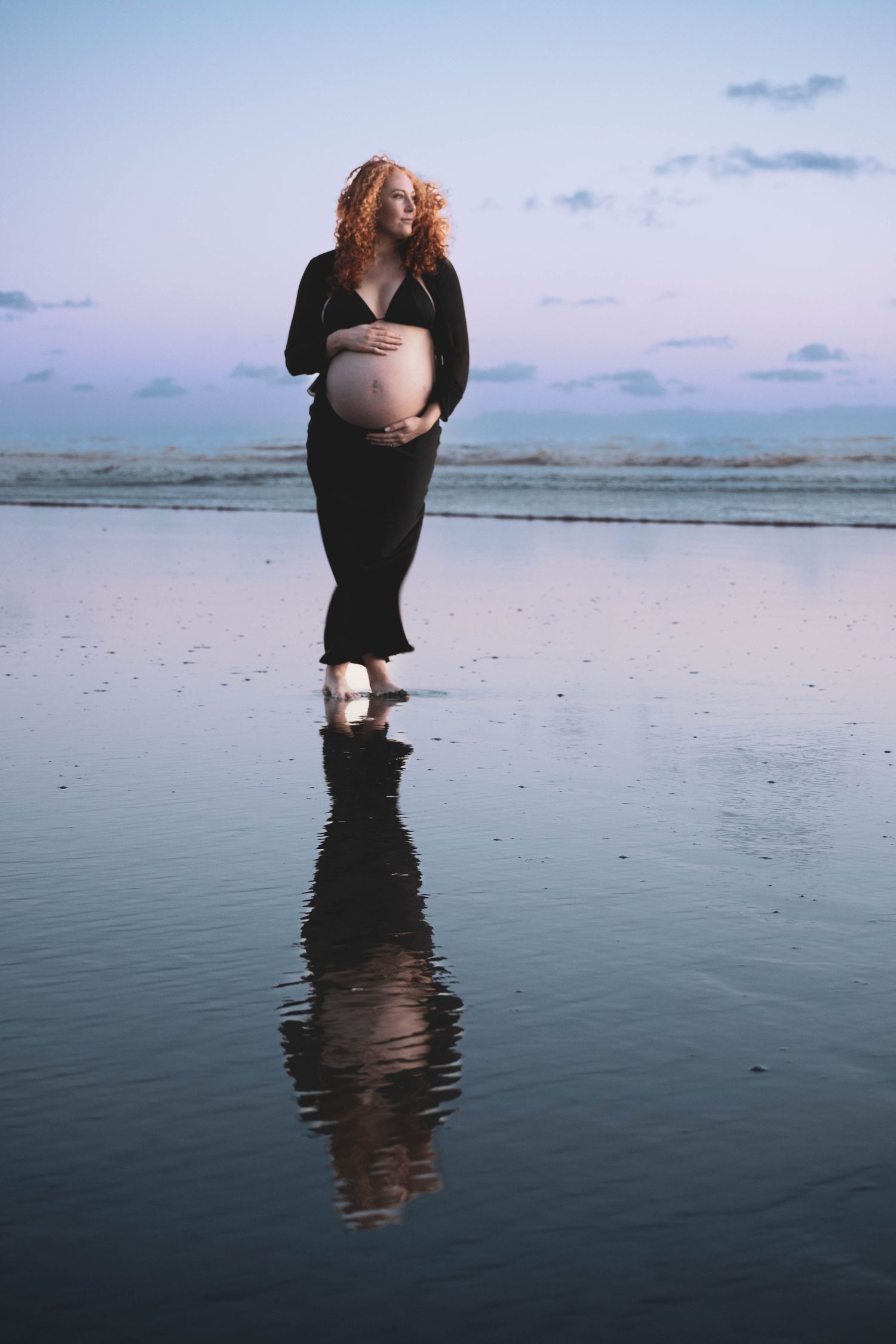 Pregnant woman in black dress cradling her belly on a reflective beach at dusk with soft purple sky.