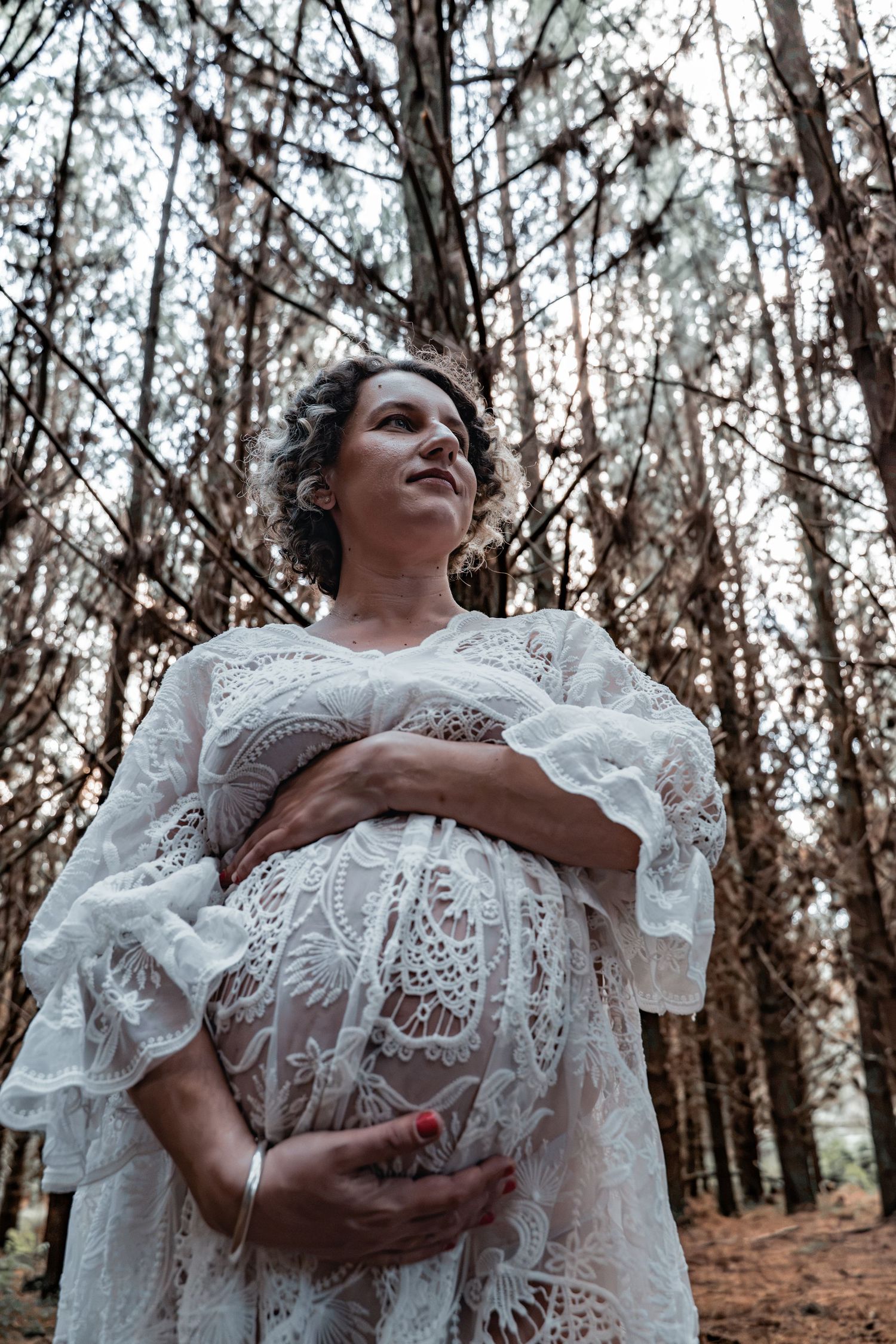 Pregnant woman in white lace dress cradling her baby bump while standing among tall trees in a forest.