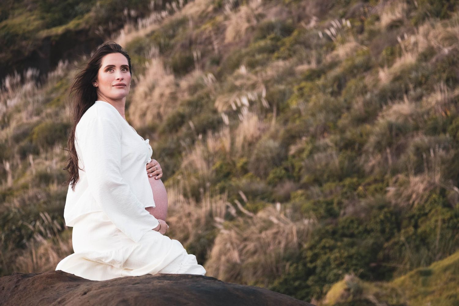 Pregnant woman in white dress sitting on rock surrounded by lush green hillside, cradling her baby bump.