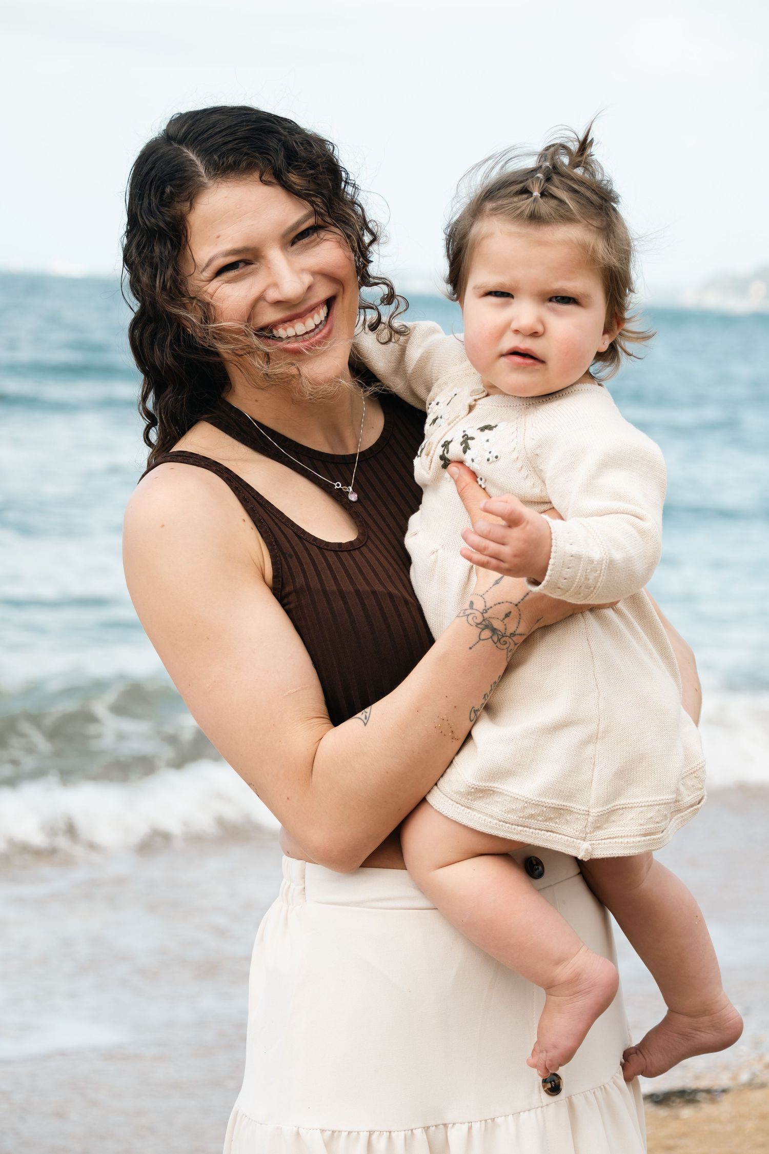 A smiling woman with curly dark hair holds a baby girl in a cream dress on a beach with ocean waves behind them.