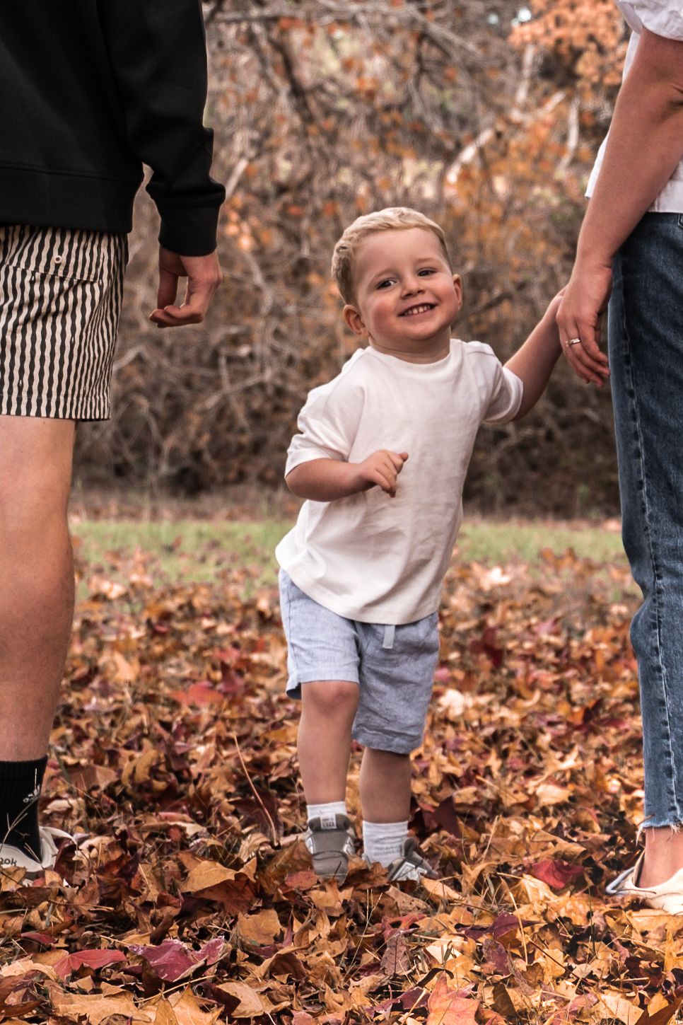 Smiling toddler boy holding hands with parents while standing in autumn leaves in a park.