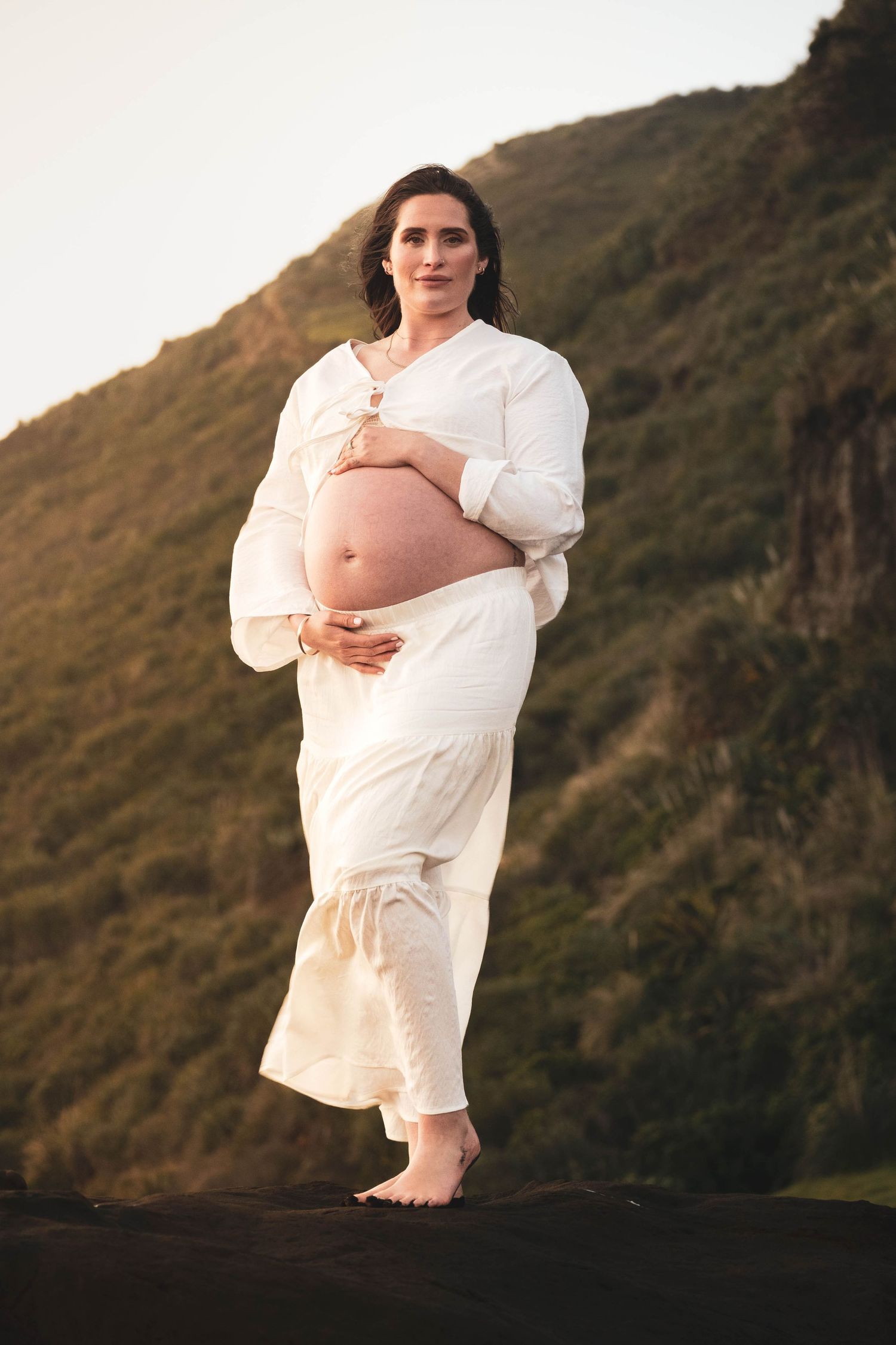 Pregnant woman in white outfit holding baby bump outdoors with green hillside backdrop at golden hour.