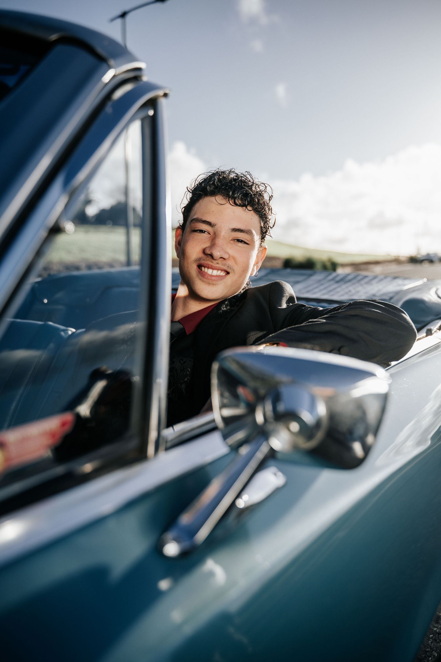 Smiling young man with curly hair leaning out of a classic blue vintage car on a sunny day.