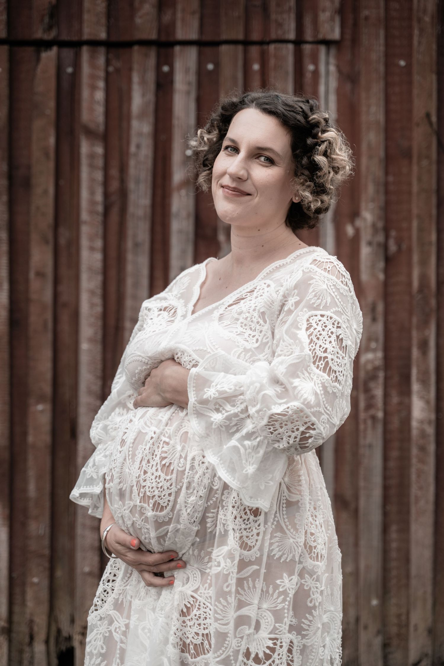Pregnant woman in white lace dress cradling baby bump, posing against rustic wooden barn wall.